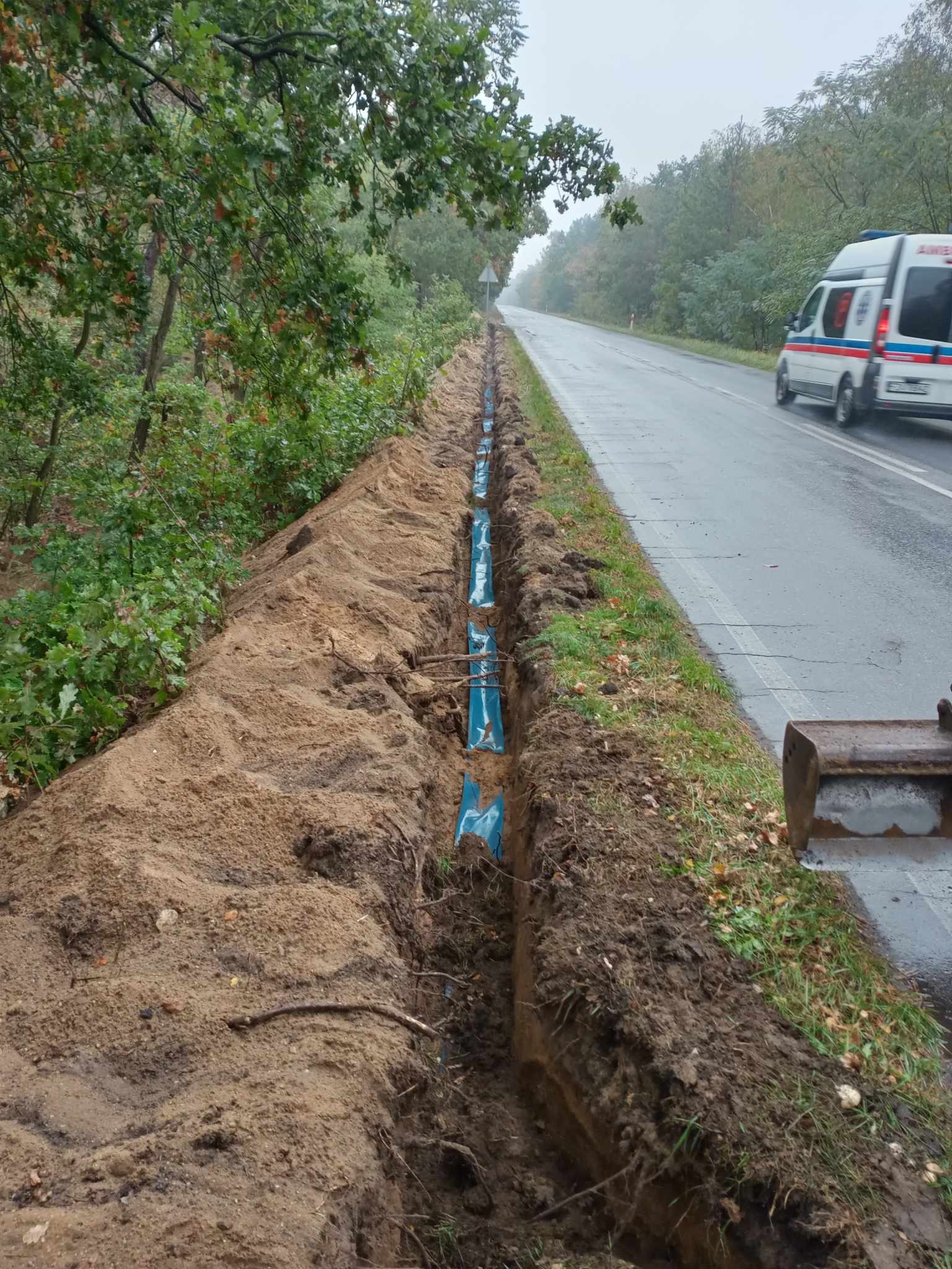 Długi wykop wzdłuż drogi asfaltowej, zabezpieczony niebieską folią, z widoczną łyżką koparki i przejeżdżającą karetką pogotowia w tle.