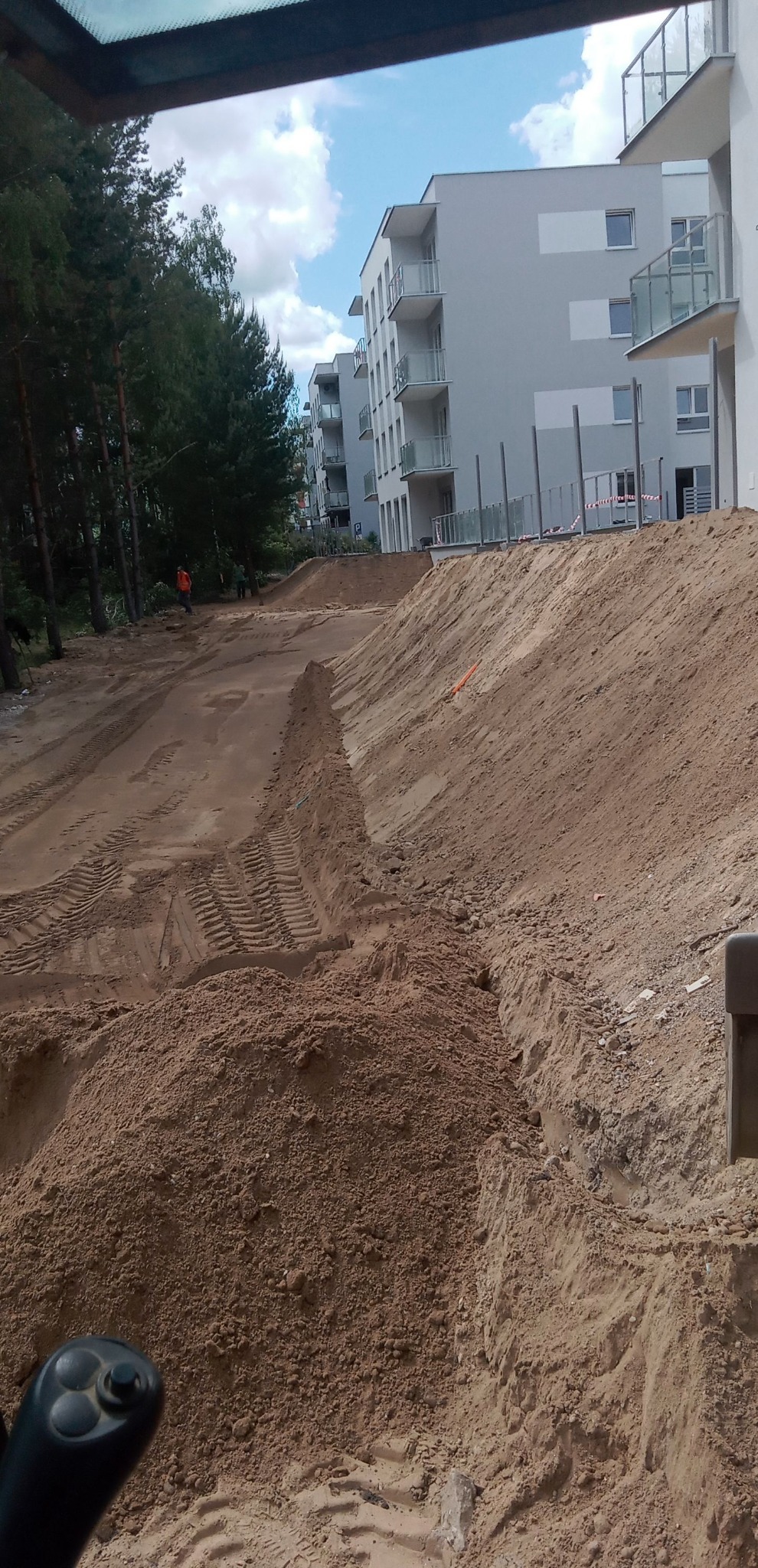Teren budowy z wyrównanym piaskiem, widok z kabiny maszyny, w tle budynki mieszkalne i drzewa.