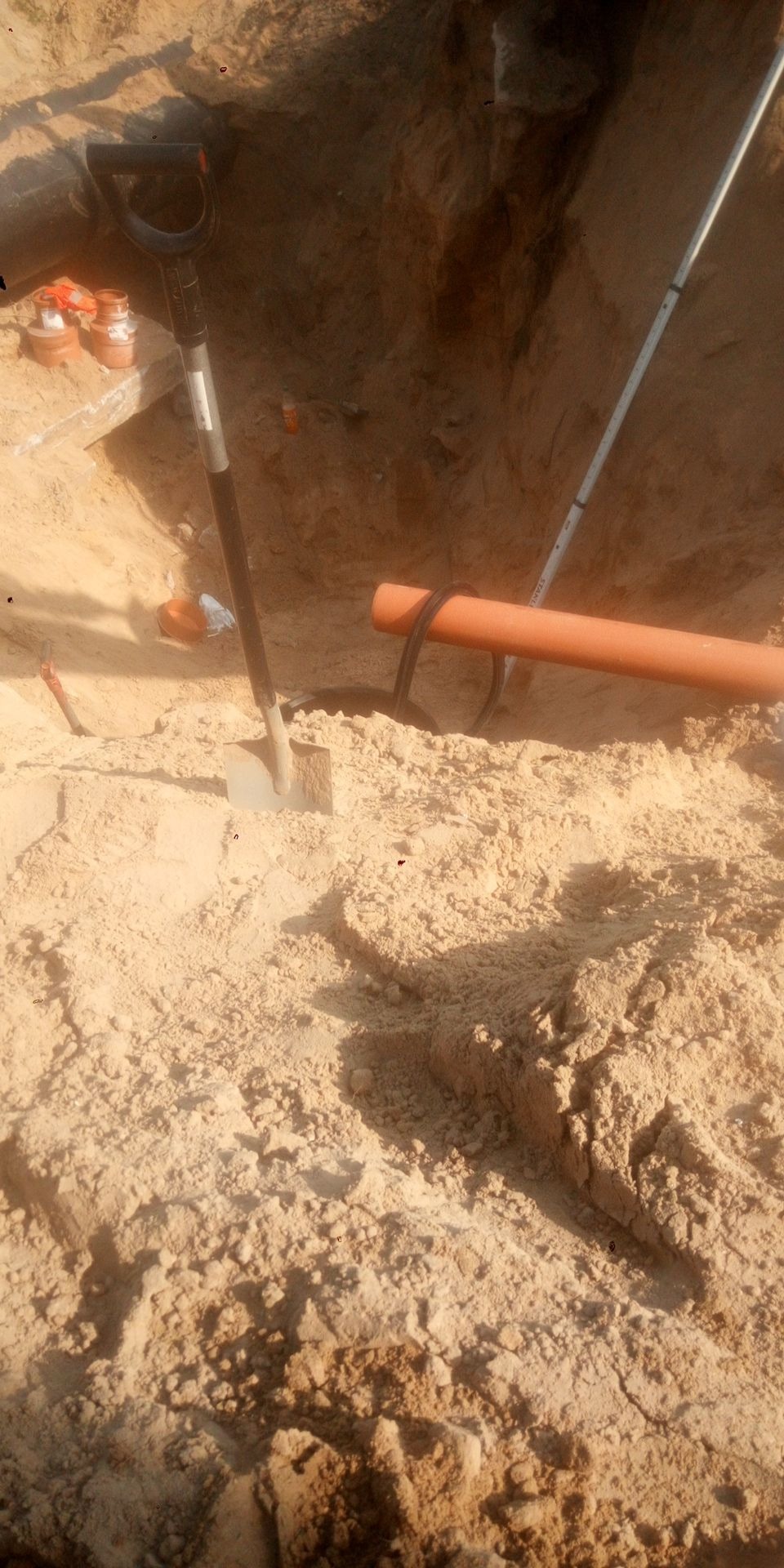 Wykop w piasku z pomarańczową rurą kanalizacyjną, łopatą i elementami instalacji, widoczne ślady pracy ziemnej.