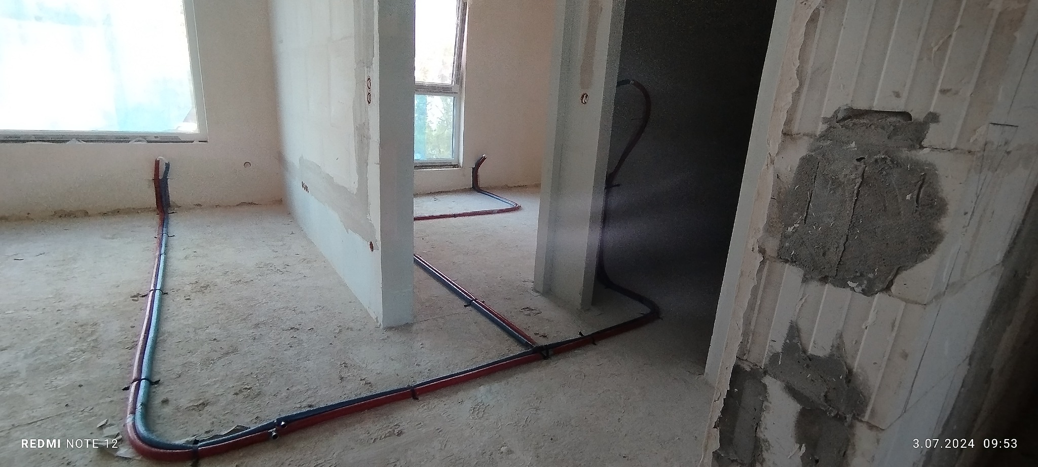 Wnętrze w trakcie prac instalacyjnych: rury grzewcze i hydrauliczne poprowadzone po podłodze w surowym stanie, widoczne okna i niedokończone ściany.