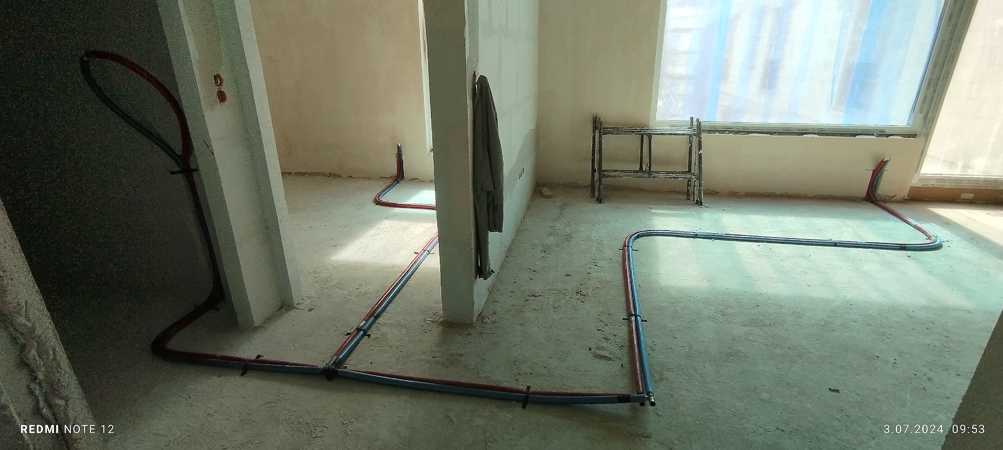 Instalacja ogrzewania podłogowego w surowym stanie, rury grzewcze ułożone na posadzce, widoczne elementy mocujące, ściany w trakcie wykończenia, w tle rusztowanie.