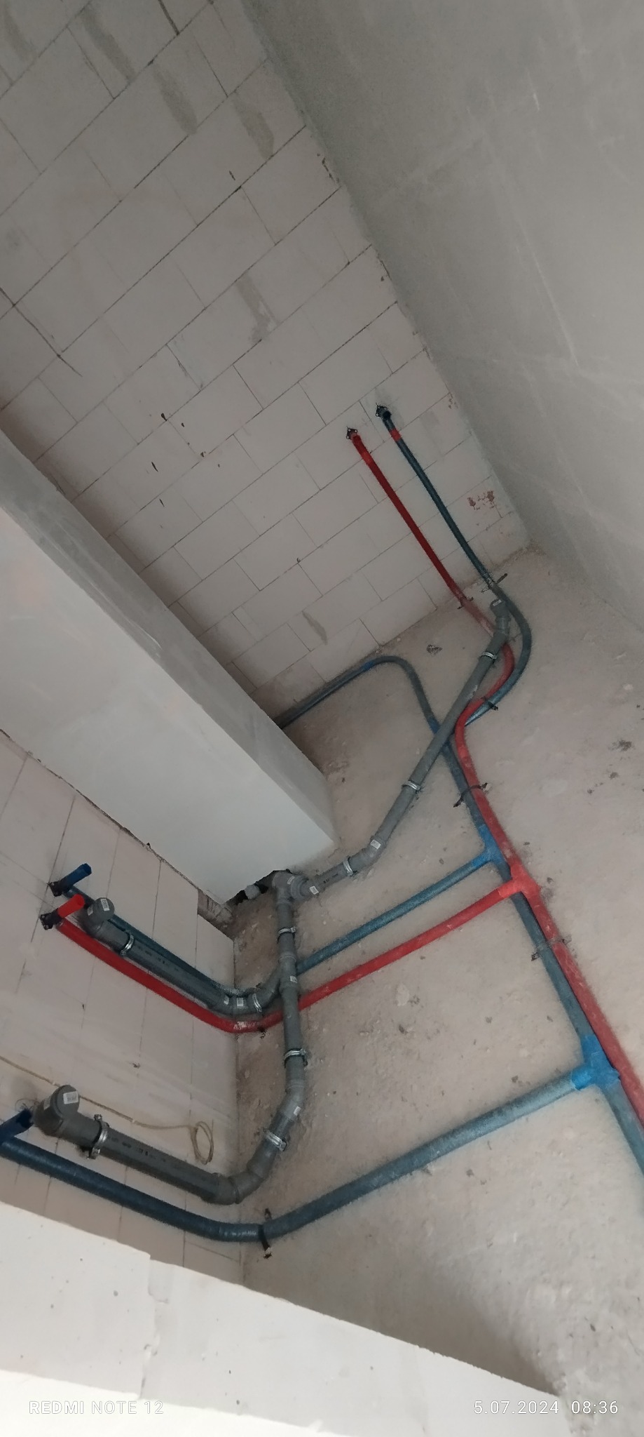 Szare rury kanalizacyjne i czerwono-niebieskie rury wodne zamontowane przy białej ścianie z pustaków i suficie w trakcie budowy.