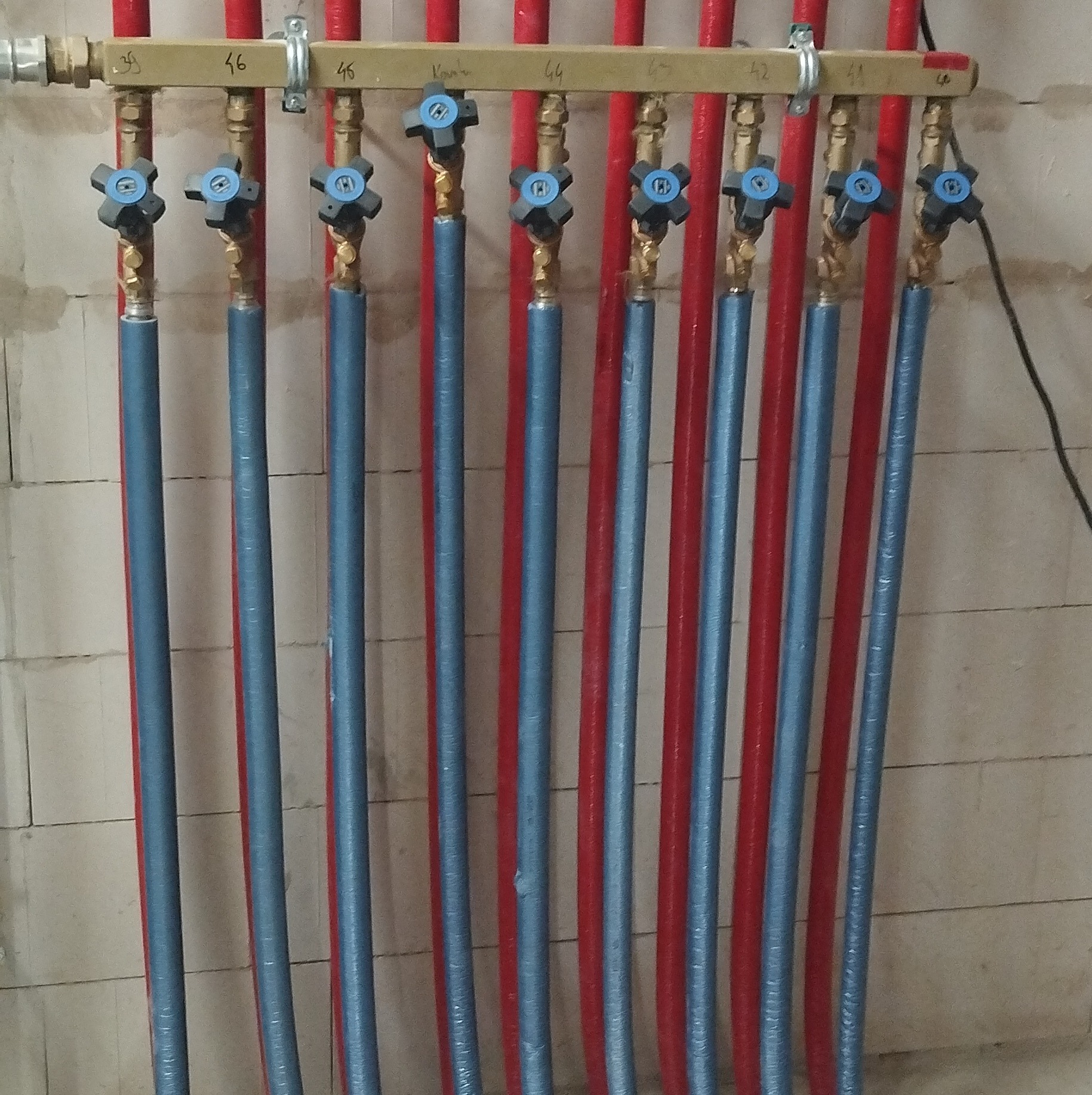 Rozdzielacz instalacji grzewczej z niebieskimi i czerwonymi rurami, zawory z niebieskimi pokrętłami, zamocowany na drewnianej listwie z numeracją, na tle ściany z bloczków.