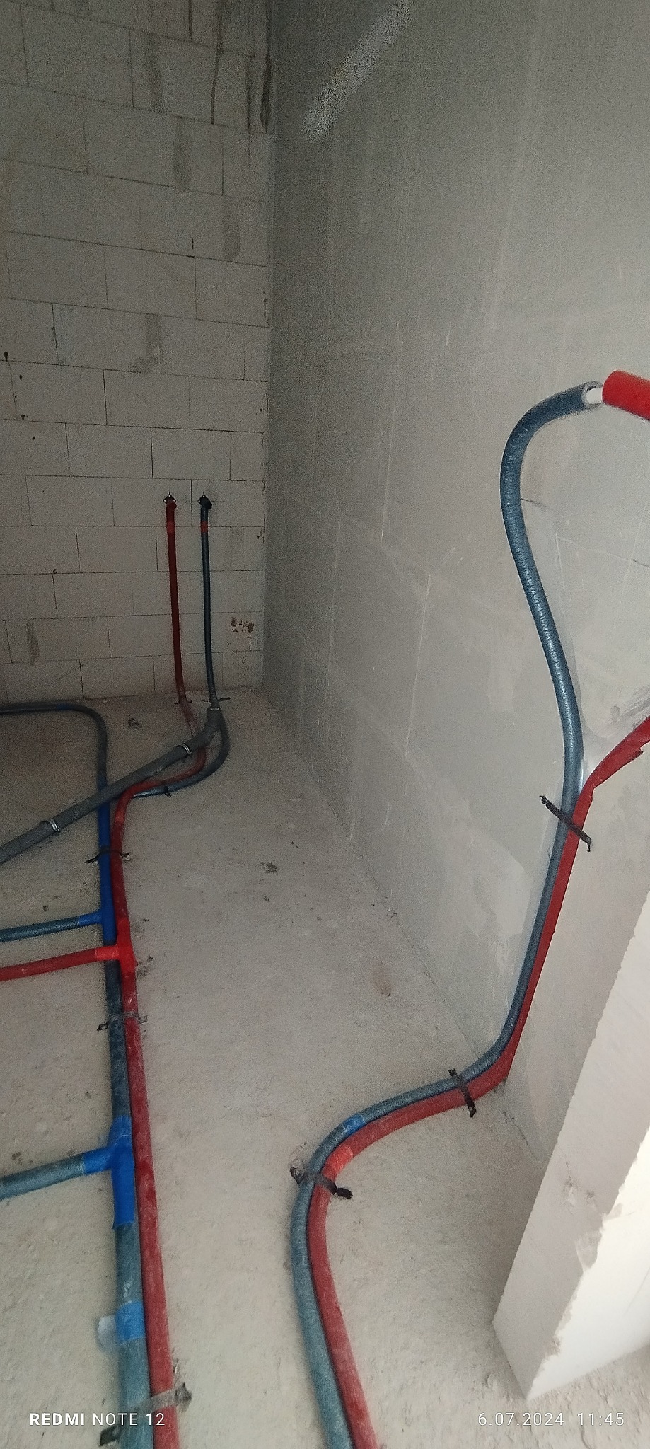 Świeżo położone, częściowo zamocowane rury instalacji hydraulicznej i grzewczej w surowym wnętrzu, z widocznymi blokami betonowymi i surową ścianą pokrytą gładzią.