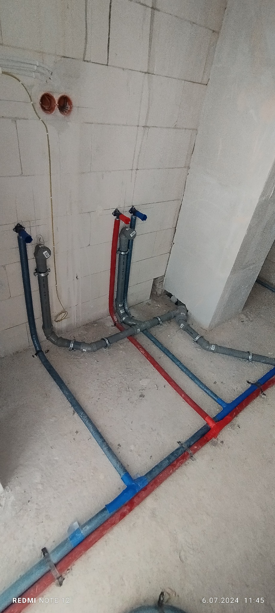 Surowe wnętrze w trakcie budowy z widocznymi rurami instalacji wodnej i kanalizacyjnej, pionowe rury z zaworami przy ścianie z betonu komórkowego, poziome rury prowadzone po podłodze, połączenia...