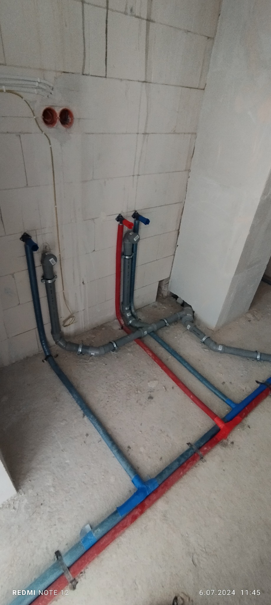 Surowe wnętrze w trakcie prac: widoczne rury instalacji wodnej (niebieskie i czerwone) prowadzone po podłodze i w górę ściany z bloczków, z zamontowanymi zaworami, oraz wystające kable elektryczne.