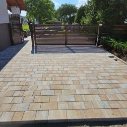 MARIO-BRUK MARIUSZ ZBORALSKI - Ułożona kostka brukowa w odcieniach beżu i szarości prowadząca do nowoczesnej, poziomej bramy wjazdowej; widoczny fragment domu po lewej i zieleń ogrodu w tle.