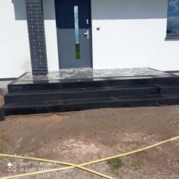 MARIO-BRUK MARIUSZ ZBORALSKI - Granitowe schody wejściowe do domu z szarymi drzwiami i ceglanym filarem, teren wokół w trakcie prac wykończeniowych, widoczny żółty wąż.