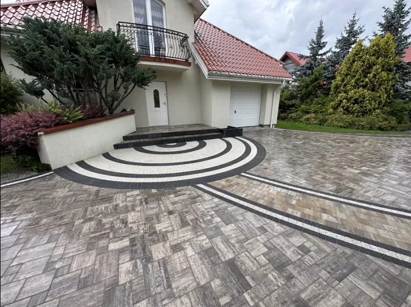 Ułożona kostka brukowa w odcieniach szarości i czerni tworząca wzór koncentrycznych okręgów przed domem z garażem, uzupełniona pasami z jasnego kruszywa.