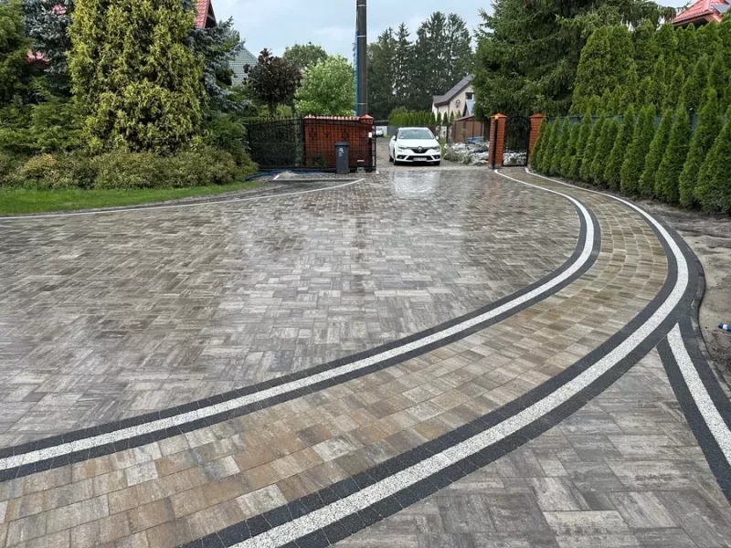 Ułożona kostka brukowa na podjeździe, wzór z dwoma białymi paskami oddzielającymi szary obszar od brązowego, widoczny samochód osobowy w tle.