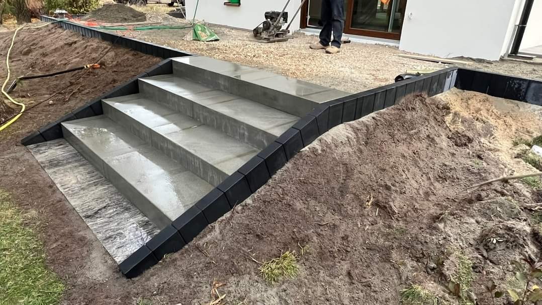 Schody z szarych płyt wykończone czarnym obrzeżem, prowadzące z podwórka do domu, w tle ubijarka wibracyjna i sterta piasku.