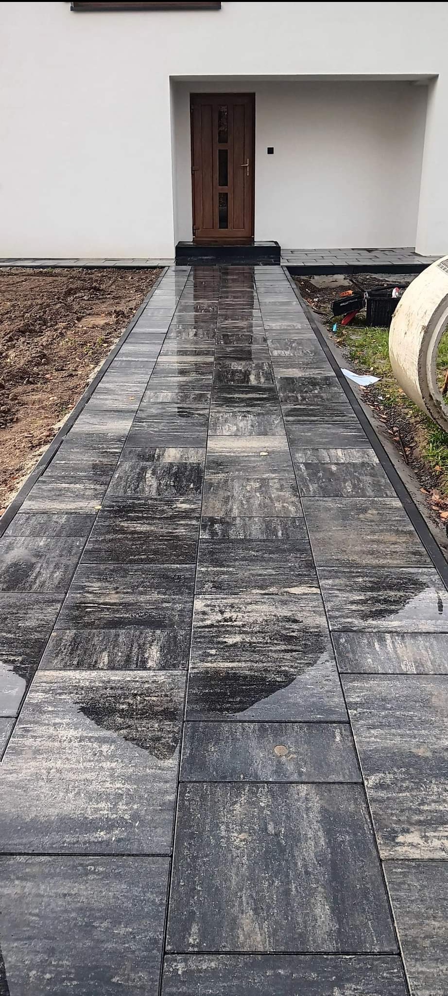 Ułożona, mokra ścieżka z grafitowej kostki brukowej prowadząca do wejścia domu z drewnianymi drzwiami, widoczne narzędzia i betonowy krąg w tle.