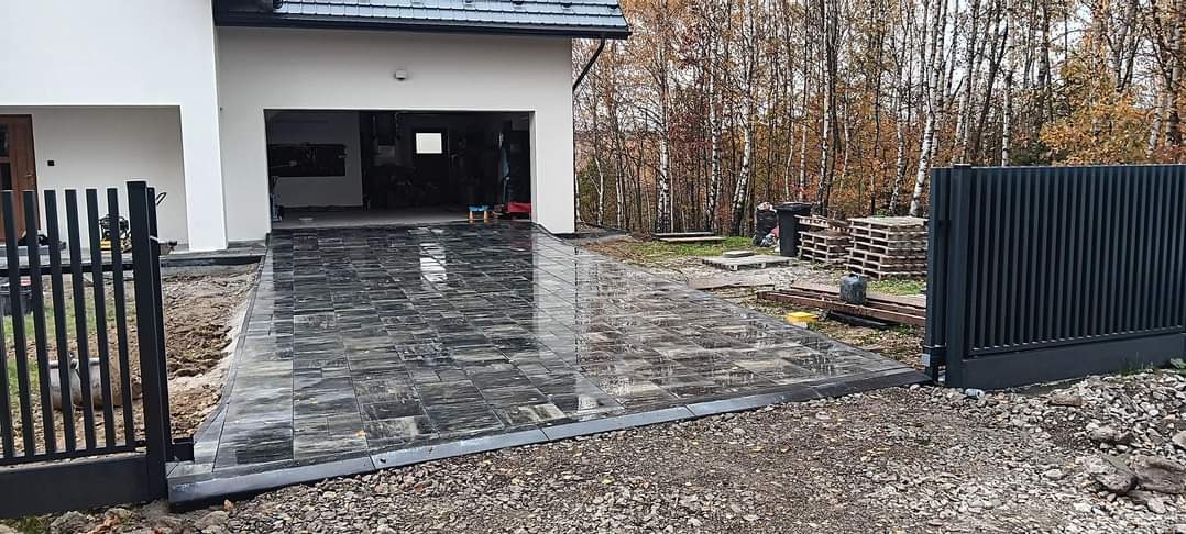 Nowo ułożona, ciemnoszara kostka na podjeździe do garażu, po deszczu, z widoczną bramą w tle i elementami krajobrazu jesiennego.