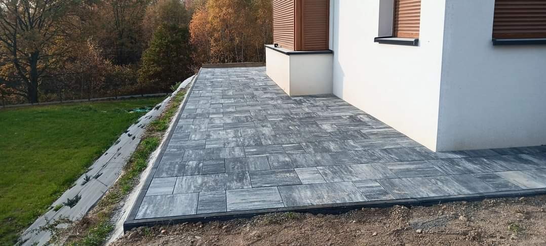 Ułożona szara kostka brukowa z czarnym obrzeżem przylegająca do białego budynku z brązowymi okiennicami, widok z perspektywy lekko z góry, trawnik na skarpie w tle.