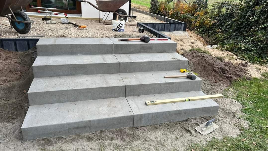 Czterostopniowe schody z szarej kostki betonowej w trakcie budowy, poziomica i młotek leżą na stopniach, widoczny fragment terenu przygotowany do dalszych prac.