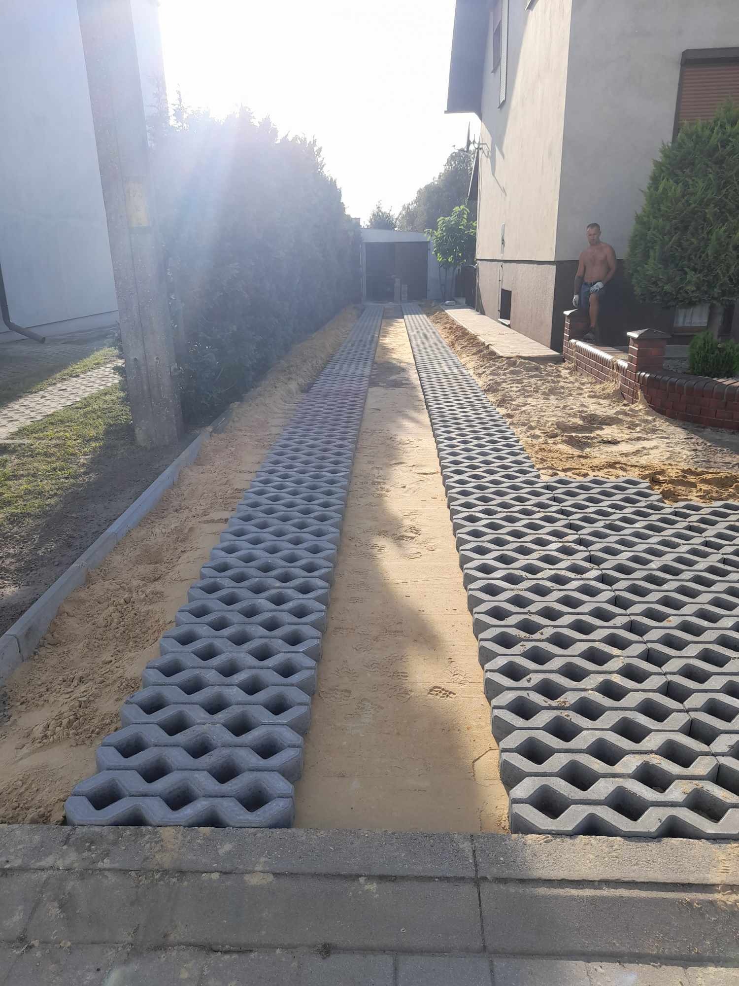 Układanie ażurowych płyt betonowych na podjeździe, widoczny piasek pomiędzy płytami, dom w tle, mężczyzna oparty o murek.