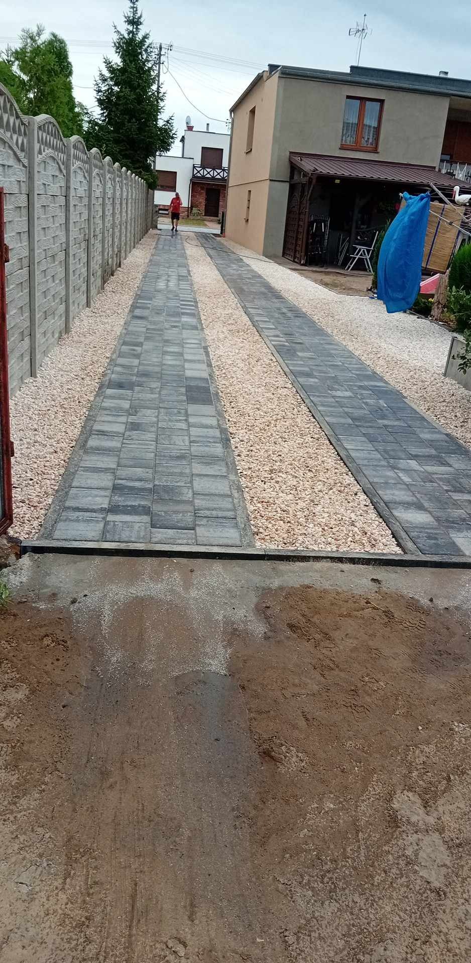Ułożona podwójna ścieżka z szarej kostki brukowej oddzielona żwirem, prowadząca do domu, widoczny betonowy płot po lewej stronie i osoba w oddali