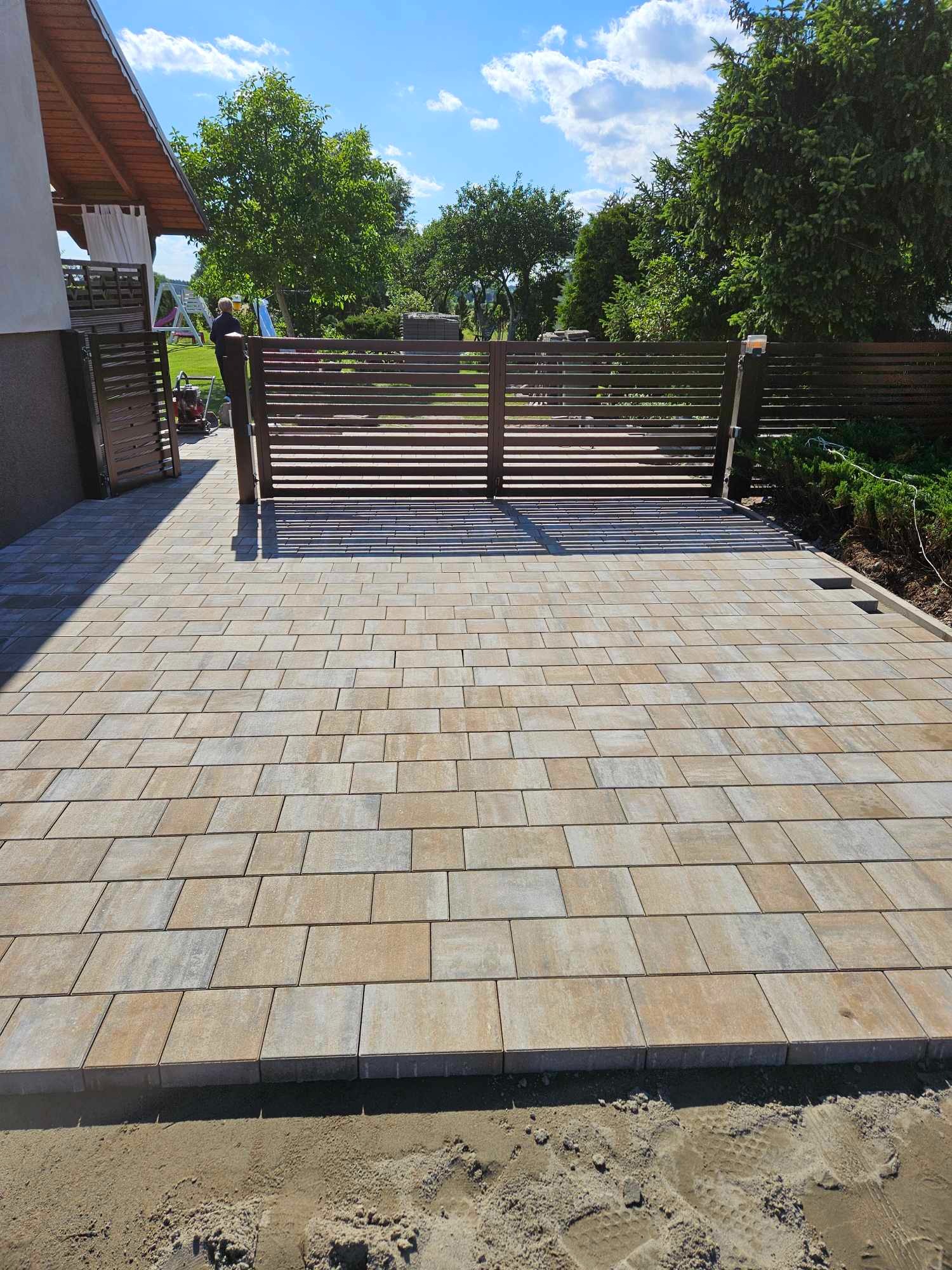 Ułożona kostka brukowa w odcieniach beżu i szarości prowadząca do nowoczesnej, poziomej bramy wjazdowej; widoczny fragment domu po lewej i zieleń ogrodu w tle.