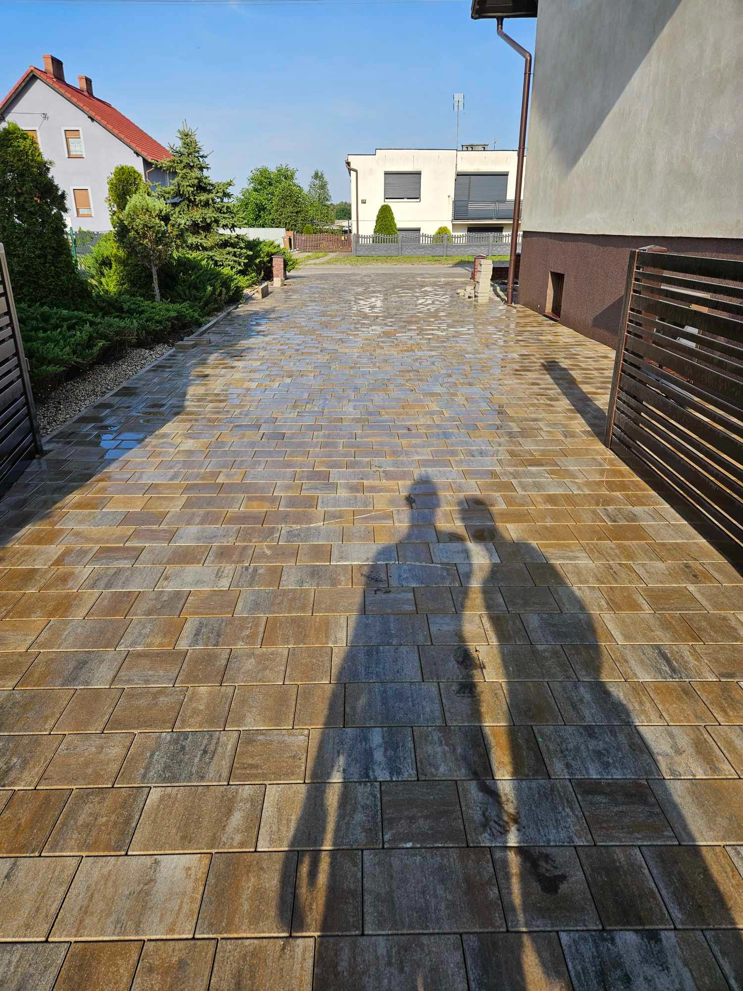 Nowa, połyskująca kostka brukowa w odcieniach brązu i szarości, ułożona na podjeździe w Kębłowie, z długimi cieniami dwóch osób wpadającymi na bruk.