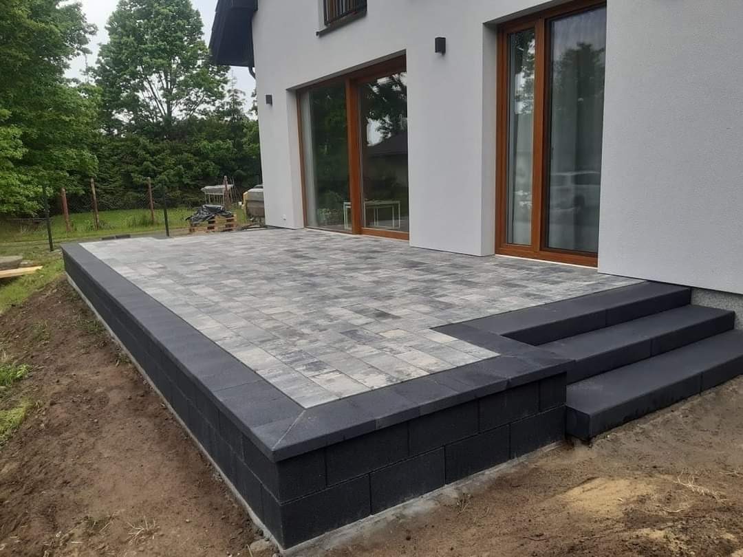Nowoczesny taras z szarej kostki brukowej ułożonej na podwyższeniu z czarnych bloczków betonowych, z trzema stopniami prowadzącymi do drzwi tarasowych domu.