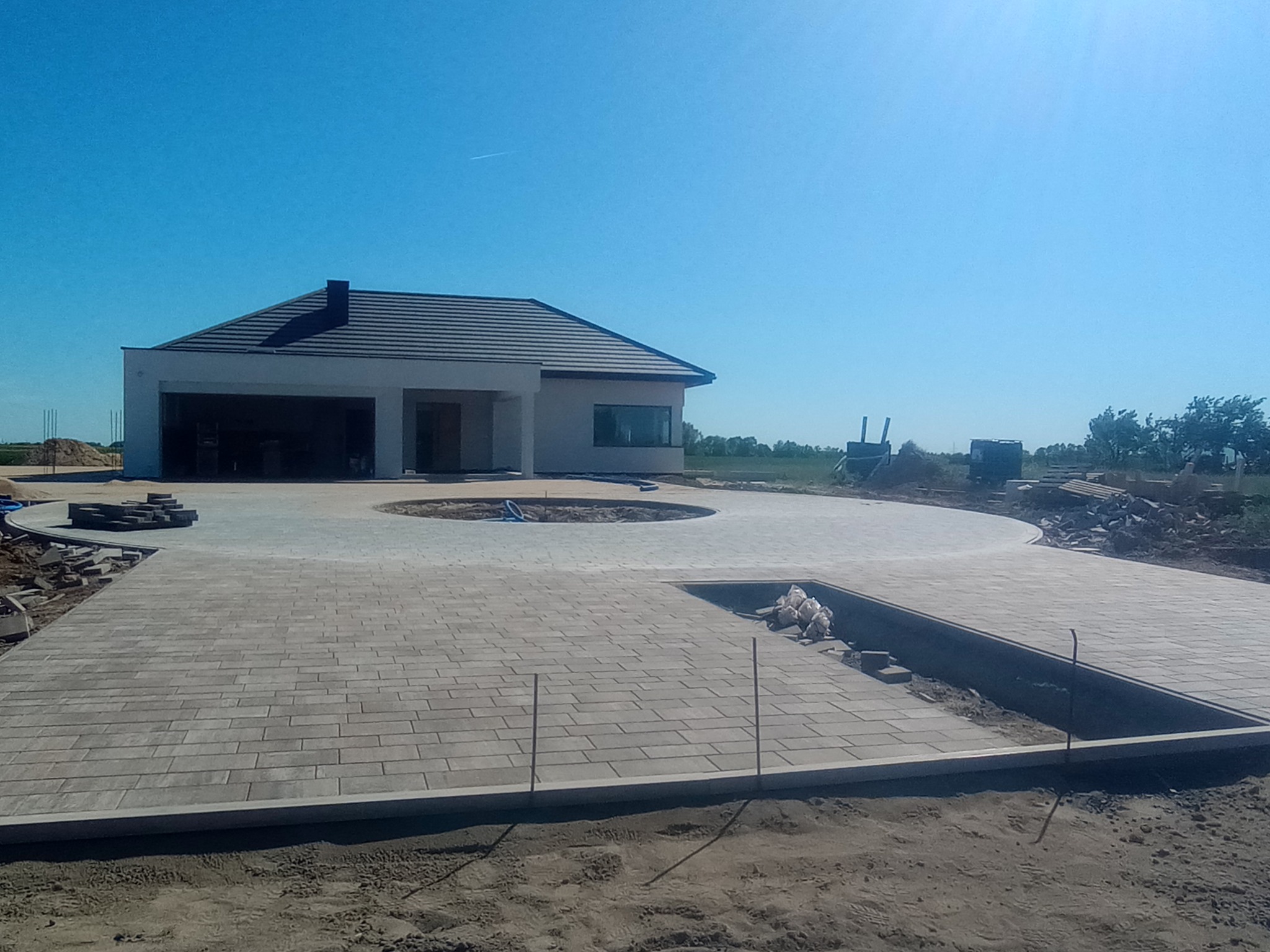Nowoczesny dom jednorodzinny z rozległym podjazdem wyłożonym szarą kostką brukową. Częściowo ułożona nawierzchnia z widocznymi elementami do dalszej pracy.
