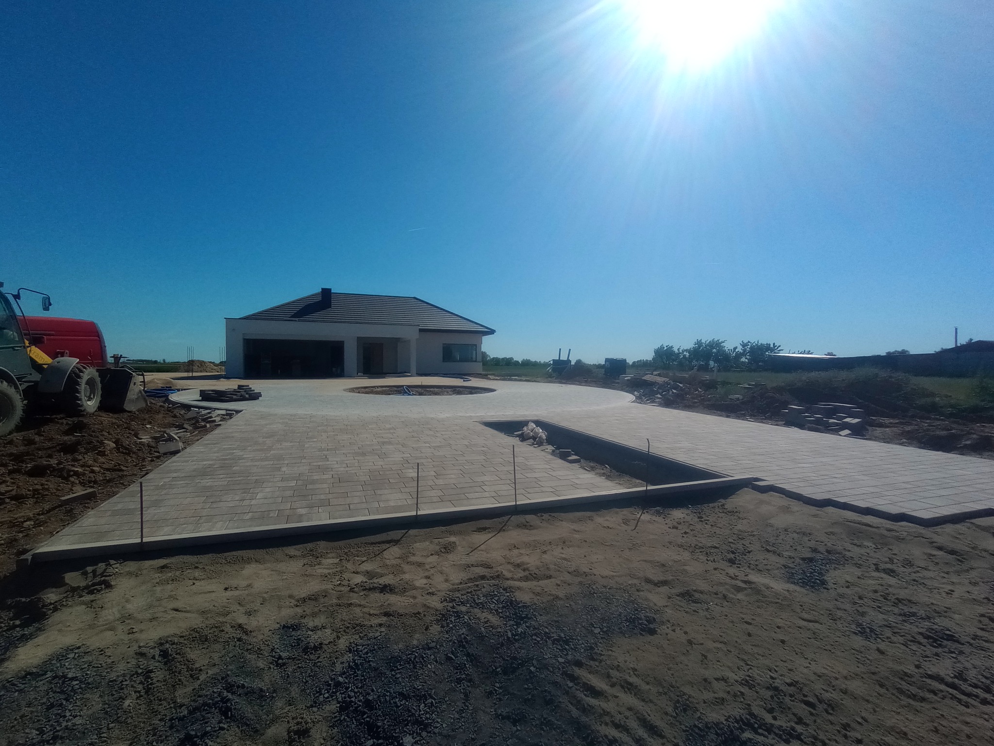 Układanie kostki brukowej na podjeździe domu z widocznym traktorem i przygotowanym terenem, jasne światło słoneczne.