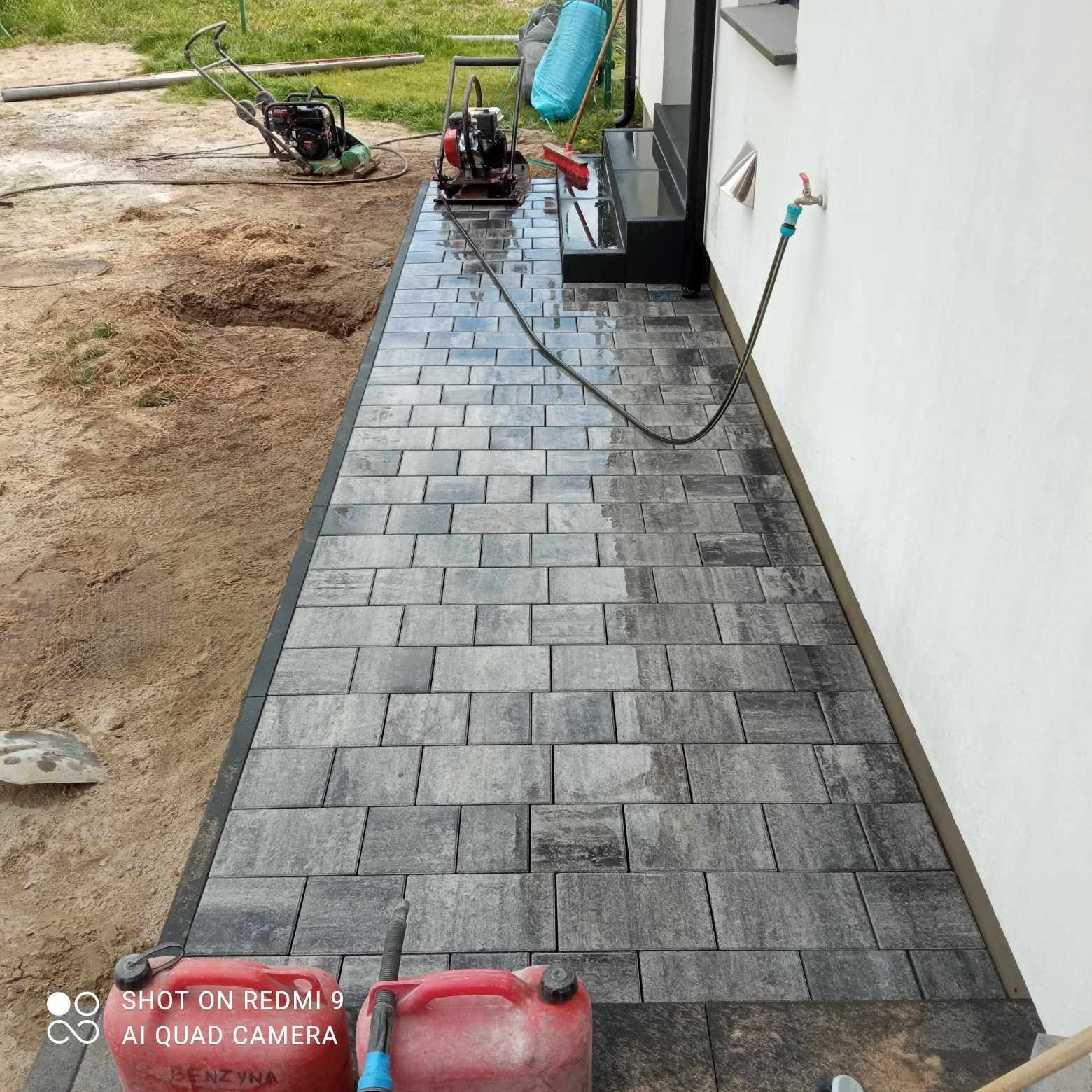 Świeżo ułożony chodnik z szarych kostek, lekko mokrych, z widocznymi zagęszczarkami, kanistrami z benzyną i wężem ogrodowym. W tle trawnik i ściana budynku.