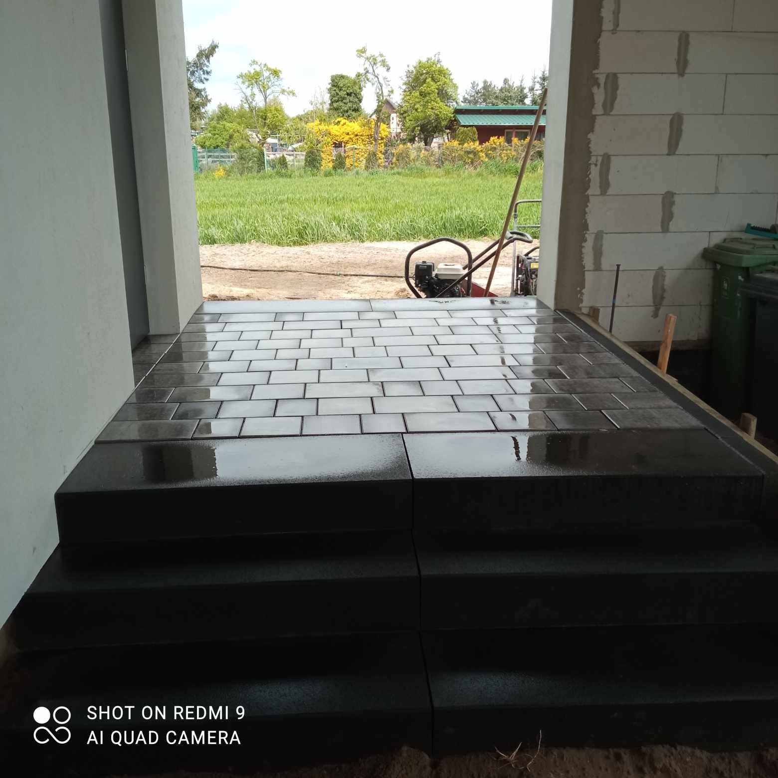 Świeżo ułożona, mokra, ciemna kostka brukowa na schodach wejściowych do budynku, z widoczną zagęszczarką w tle i surowymi ścianami budynku.