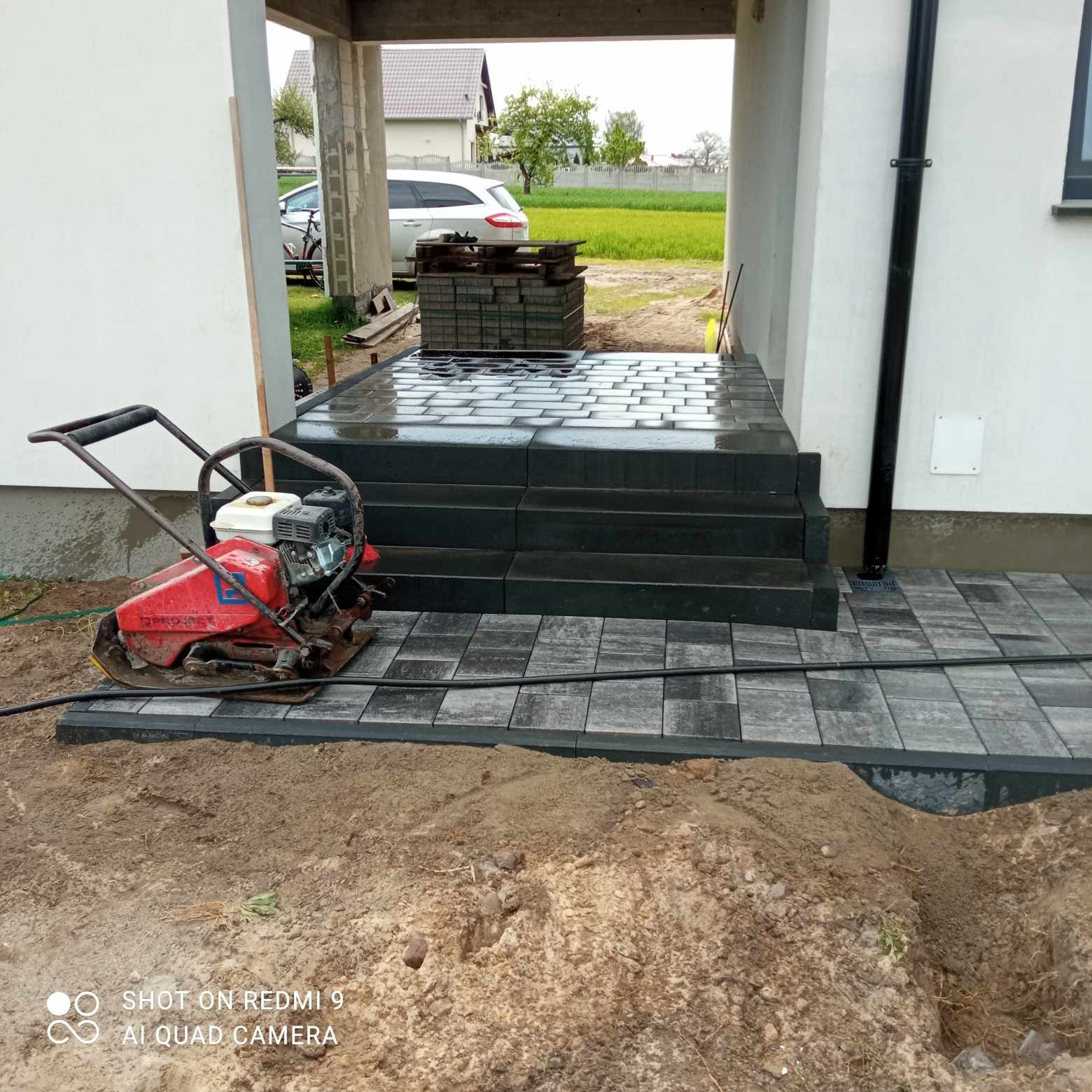 Częściowo ułożona kostka brukowa w odcieniach szarości prowadząca do wejścia do domu, z widoczną ubijarką wibrującą i stosem kostki w tle.
