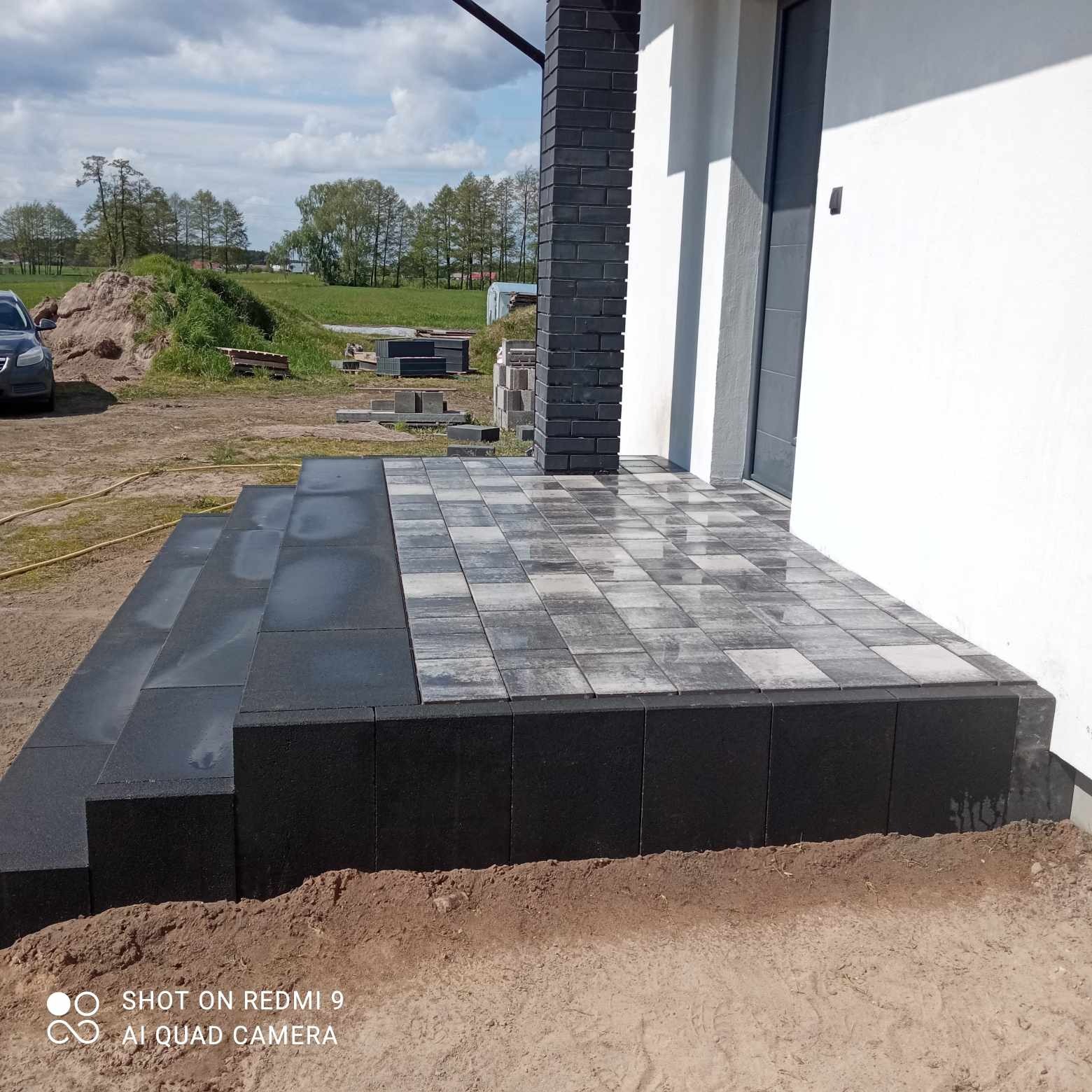 Nowo ułożone schody wejściowe i taras z kostki brukowej w odcieniach szarości, z czarnym obrzeżem i filarem z cegieł, na tle budynku w trakcie wykończenia i krajobrazu.