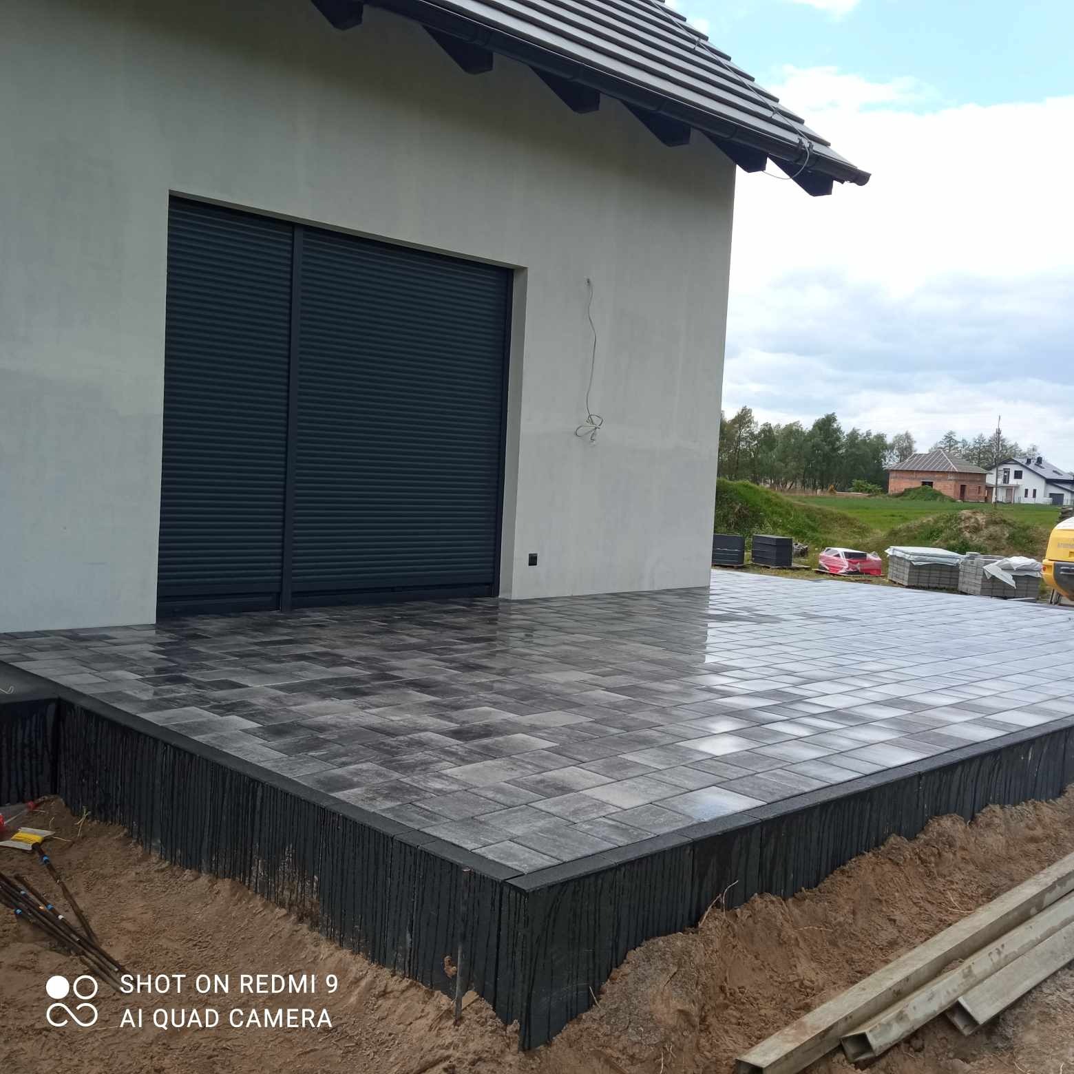 Nowo ułożona, mokra kostka brukowa w odcieniach szarości na tarasie domu z ciemnymi roletami, widoczne przygotowane elementy konstrukcyjne i odsłonięta ziemia wokół tarasu, sugerująca trwające...