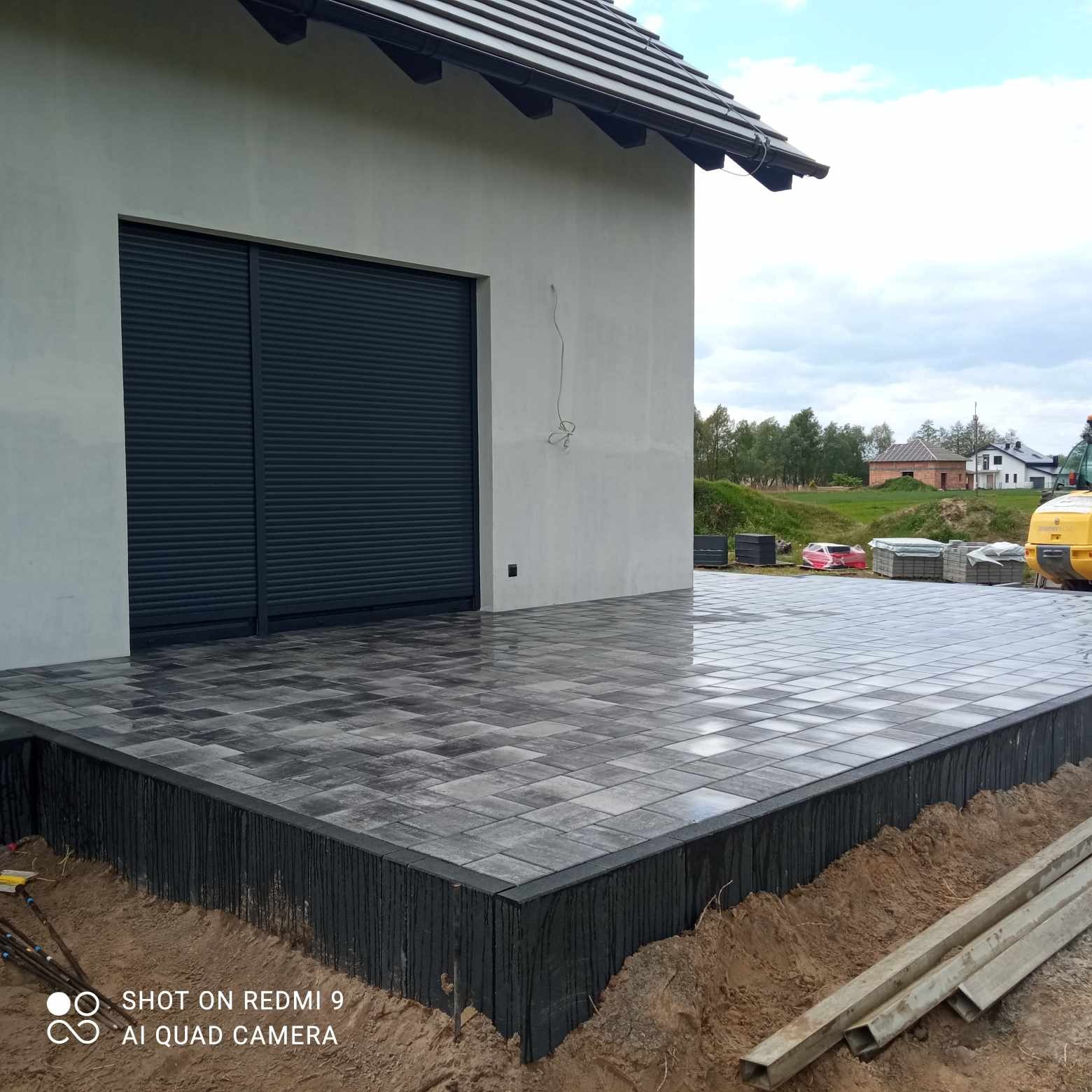 Świeżo ułożony taras z szarej kostki brukowej, przylegający do budynku z ciemnymi roletami, widoczne przygotowania do obsypania ziemią i wyrównania terenu, w tle lekki sprzęt budowlany.