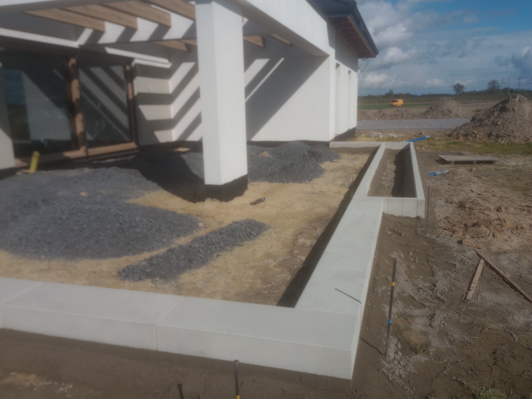 Budowa domu w trakcie realizacji: przygotowany teren pod taras z wysypanym kamieniem, obok fundament pod wylewkę, w tle hałdy ziemi i żółty samochód dostawczy.