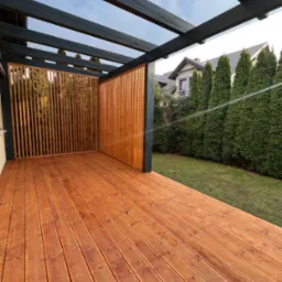 Drewniany taras z pergolą z pionowymi lamelami, widok z góry na deski, w tle zieleń i dom. Nowoczesna konstrukcja z elementami naturalnego drewna.