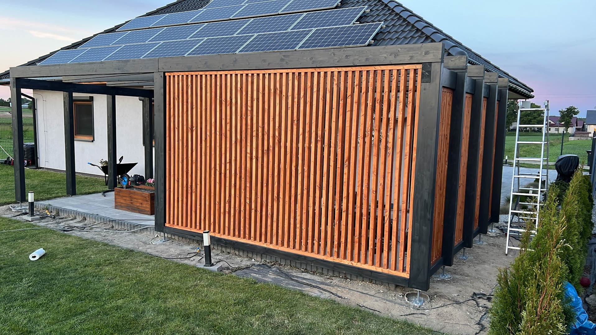 Nowoczesna pergola z drewnianymi lamelami w kolorze miodowym i czarną konstrukcją, częściowo osłonięta, na tle domu z panelami słonecznymi na dachu, widoczna drabina i elementy wykończeniowe.