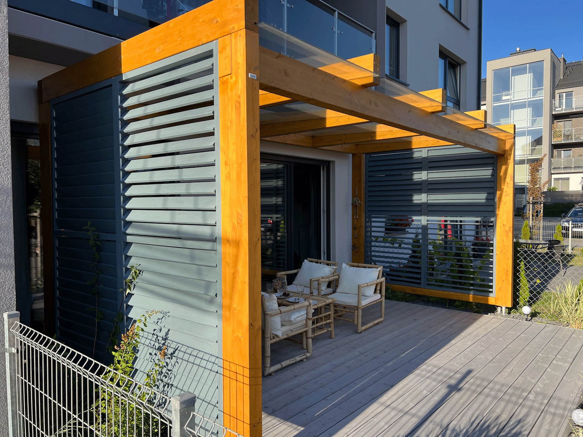 Nowoczesna pergola z drewnianą konstrukcją i szklanym zadaszeniem, połączona z tarasem z desek kompozytowych, z meblami ogrodowymi z bambusa i regulowanymi żaluzjami w kolorze szarym, na tle...