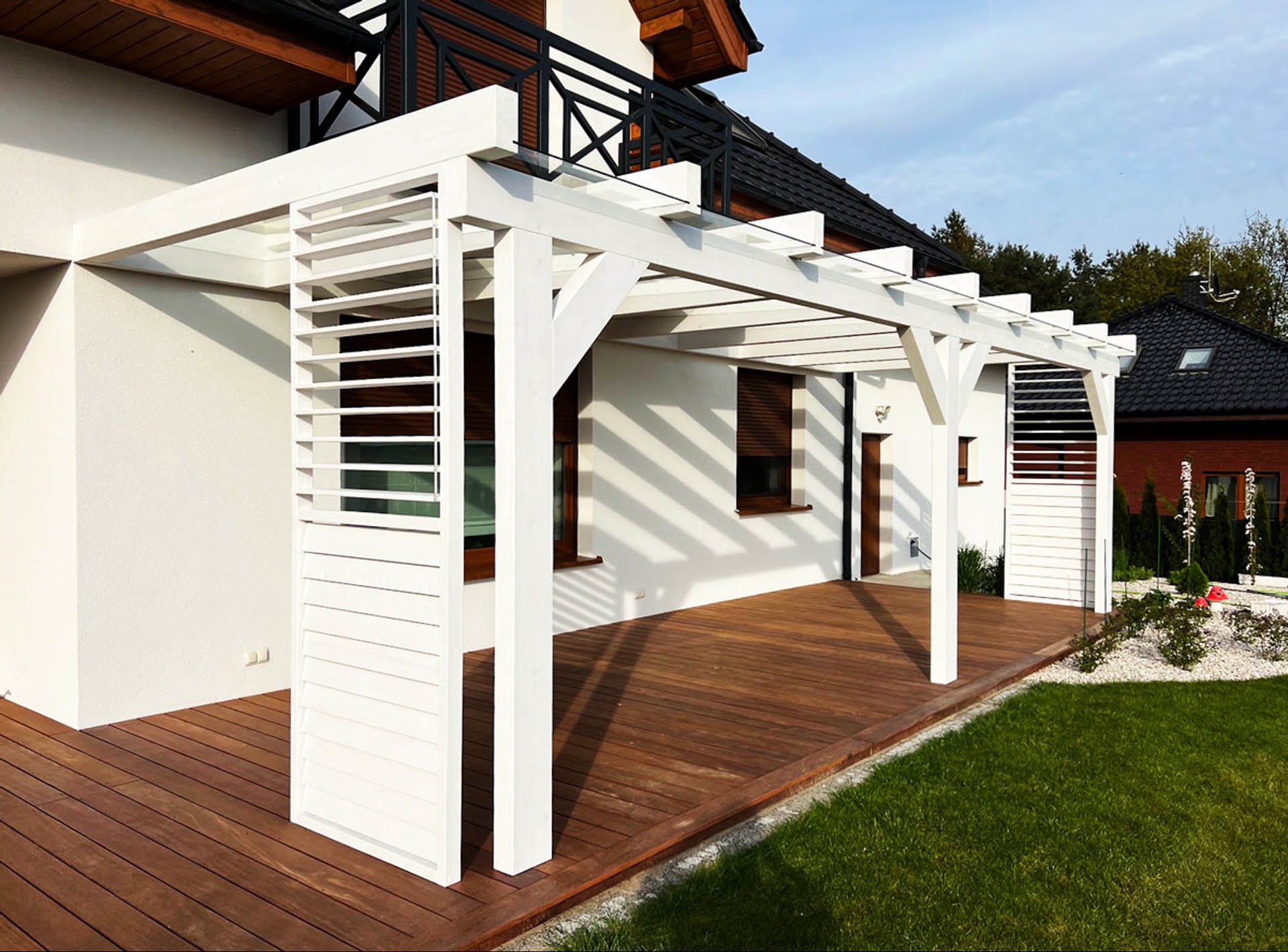 Biała pergola z lamelami na drewnianym tarasie przylegającym do nowoczesnego domu z ciemną stolarką okienną i balustradą balkonową.