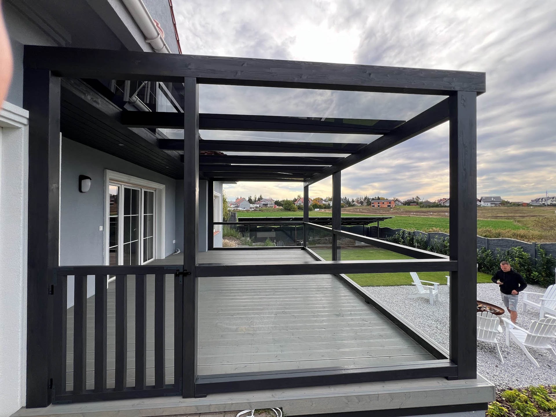 Zadaszona, grafitowa konstrukcja z drewnianymi słupami i przezroczystym dachem, przylegająca do domu z szarymi ścianami, z widocznym fragmentem tarasu z desek i białymi krzesłami ogrodowymi...