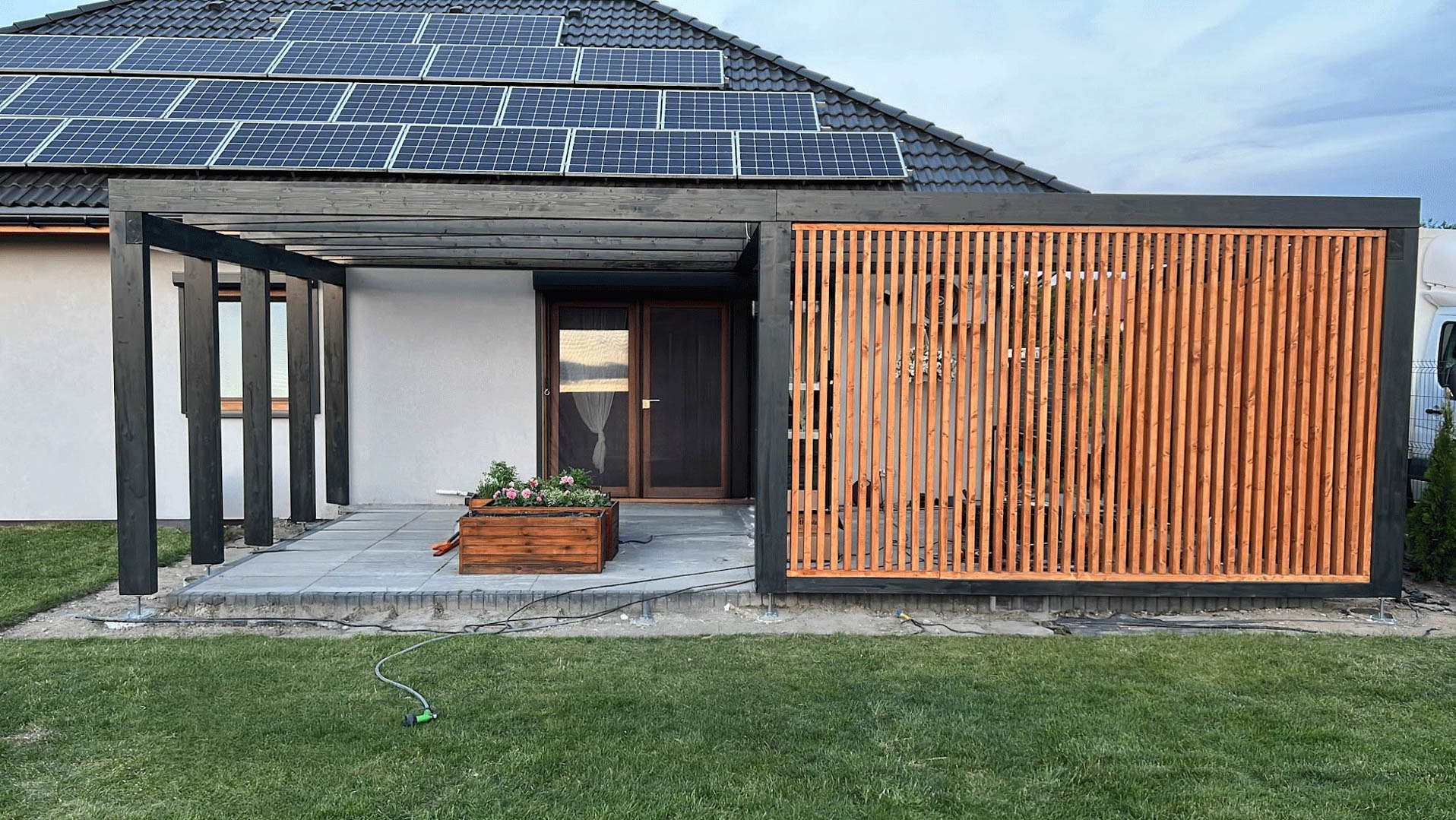Nowoczesna pergola z drewnianymi lamelami w kolorze naturalnego drewna, połączona z czarną konstrukcją, przylegająca do białego budynku z panelami słonecznymi na dachu. Na pierwszym planie kostka...