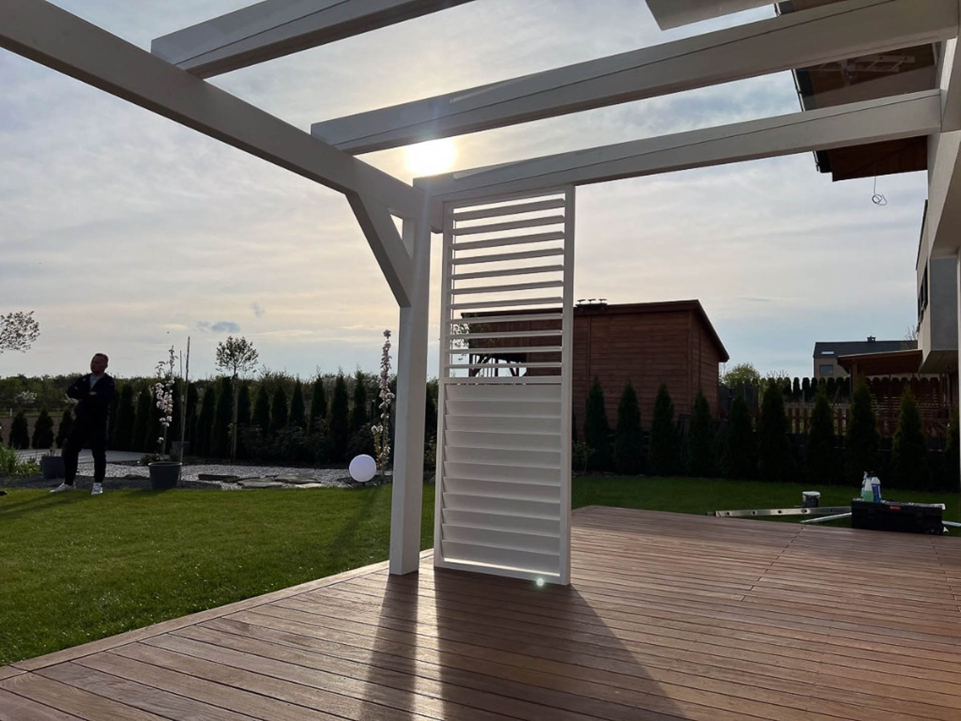 Nowoczesna pergola z białymi lamelami i drewnianym tarasem; w tle zadbany ogród z tujami i mężczyzna w garniturze.