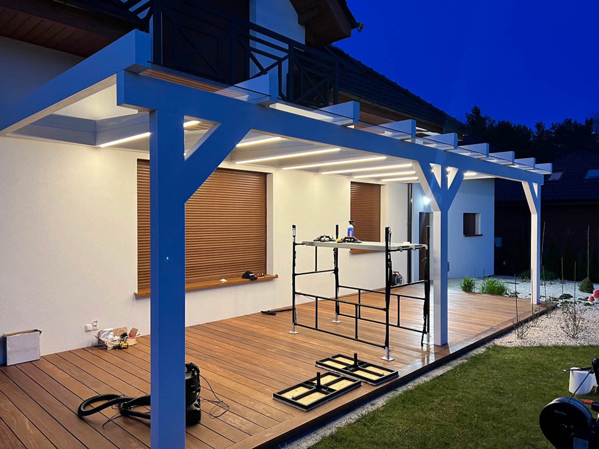 Montaż nowoczesnej, białej pergoli z oświetleniem LED nad drewnianym tarasem w Tarnowie Podgórnym, widoczne rusztowanie i narzędzia, praca w toku.