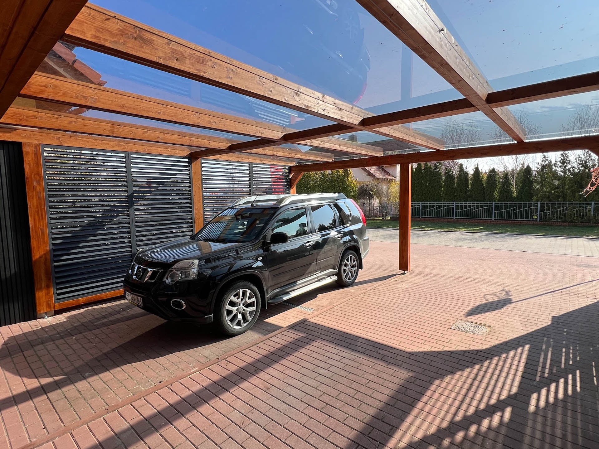 Drewniana pergola z przeszklonym dachem nad samochodem na podjeździe z czerwonej kostki brukowej, w tle widoczny ogród.