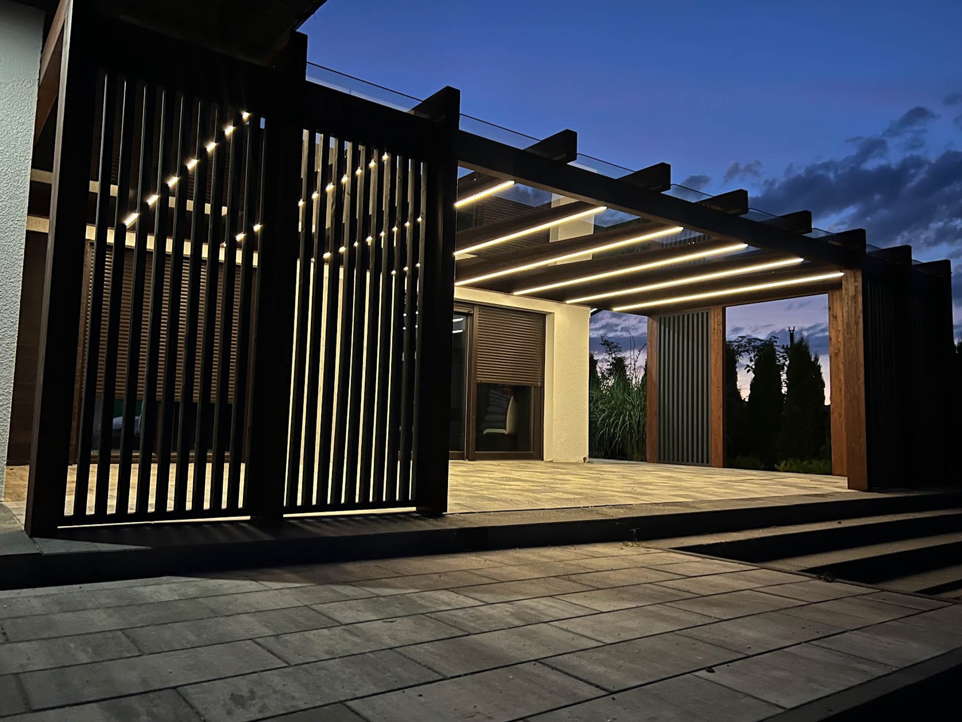 Nowoczesna pergola z drewnianą konstrukcją i oświetleniem LED, widok na taras z kostki brukowej o zmierzchu.