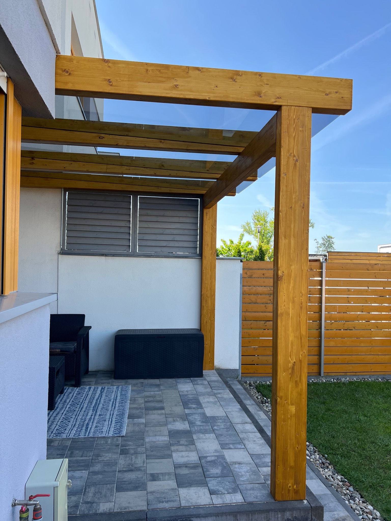 Nowoczesna, drewniana pergola z przezroczystym zadaszeniem, oświetlona słońcem, z meblami ogrodowymi i kostką brukową w tle. Widok z niskiego kąta.