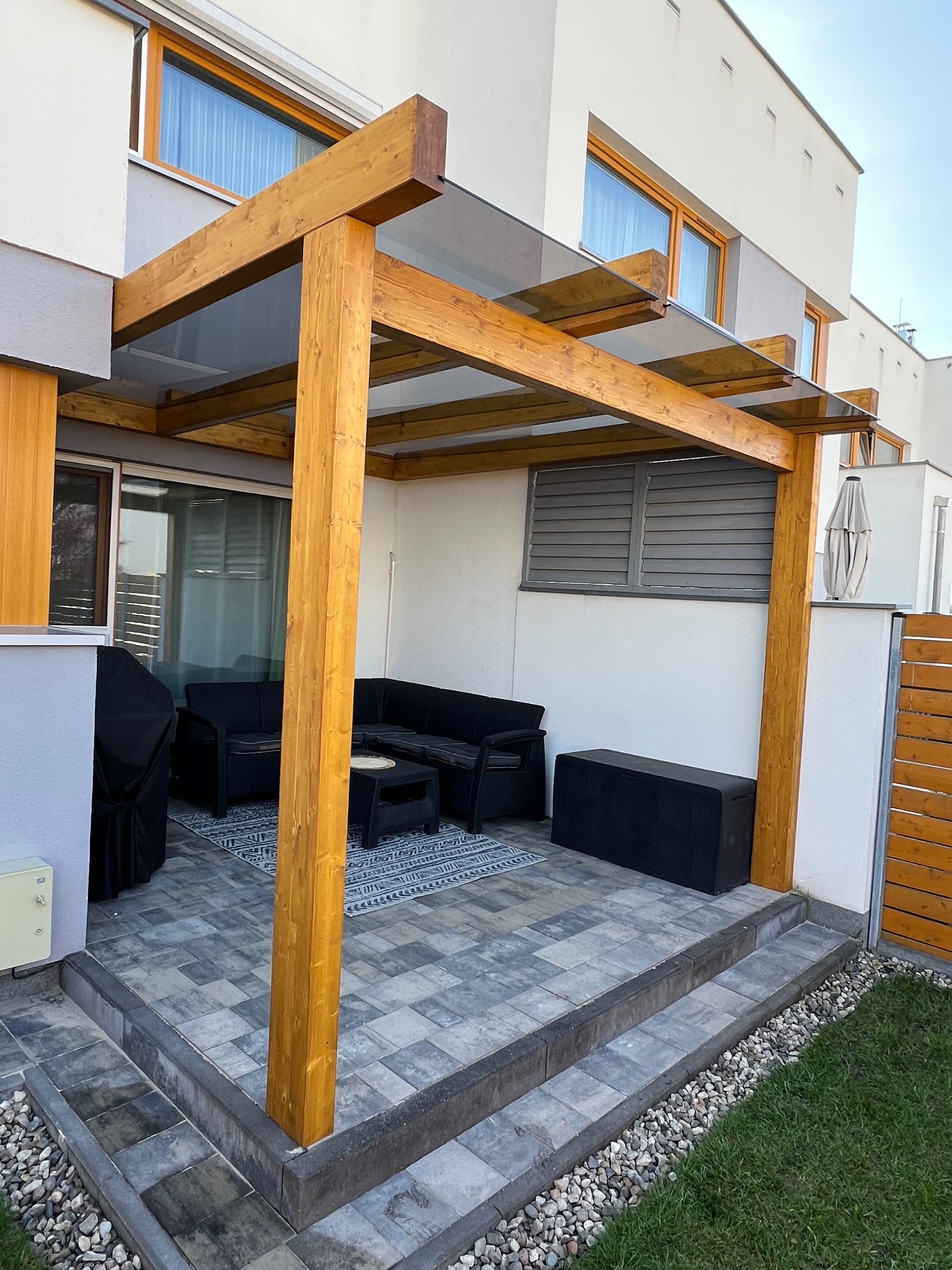 Drewniana pergola z przezroczystym zadaszeniem, meble ogrodowe na wyłożonym płytkami tarasie, nowoczesny dom w tle, błękitne niebo w górnej części kadru.