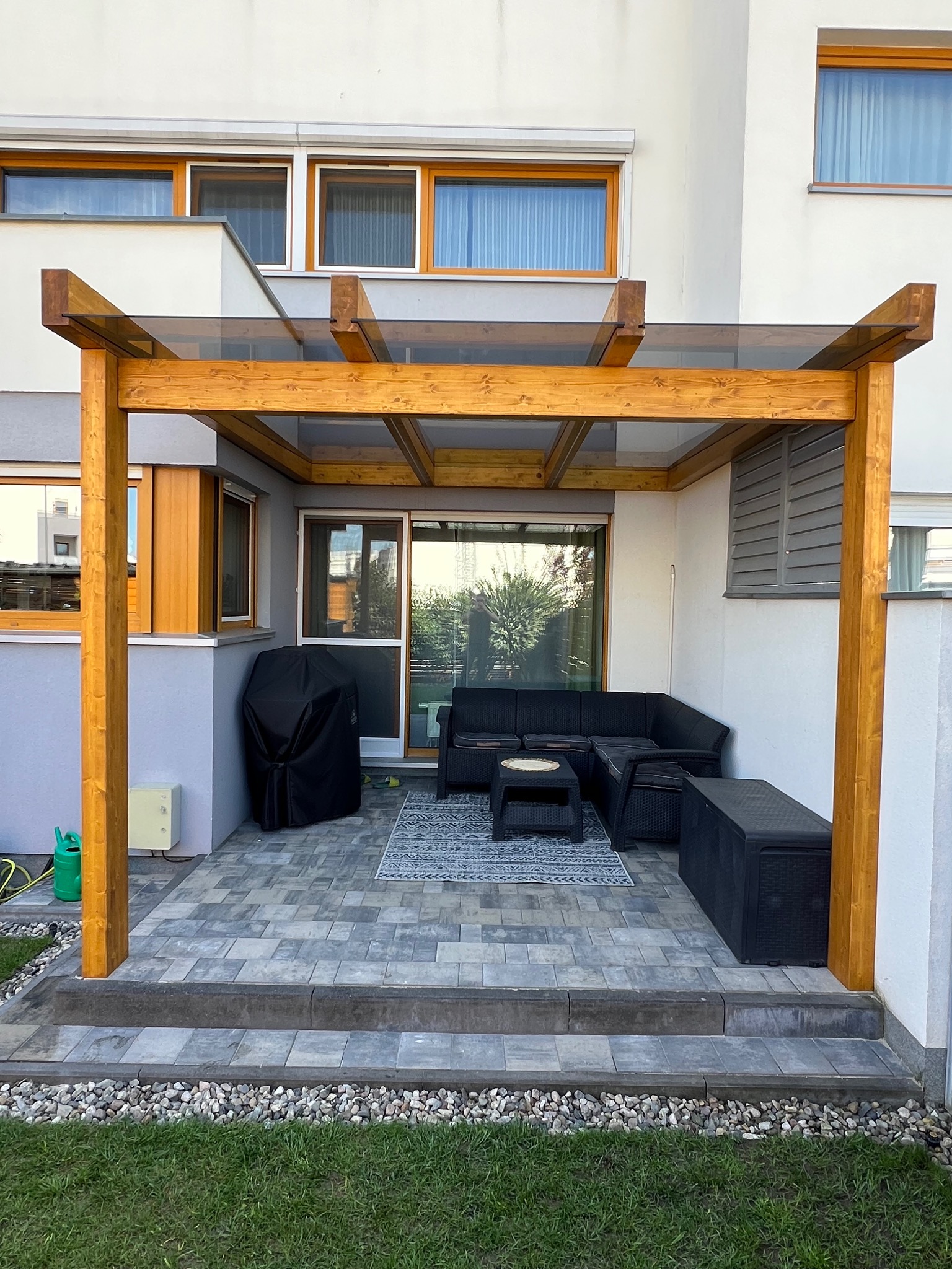 Drewniana pergola z przezroczystym zadaszeniem, pod nią meble ogrodowe i grill. Wyjście na taras z kostki brukowej, obok trawnik.