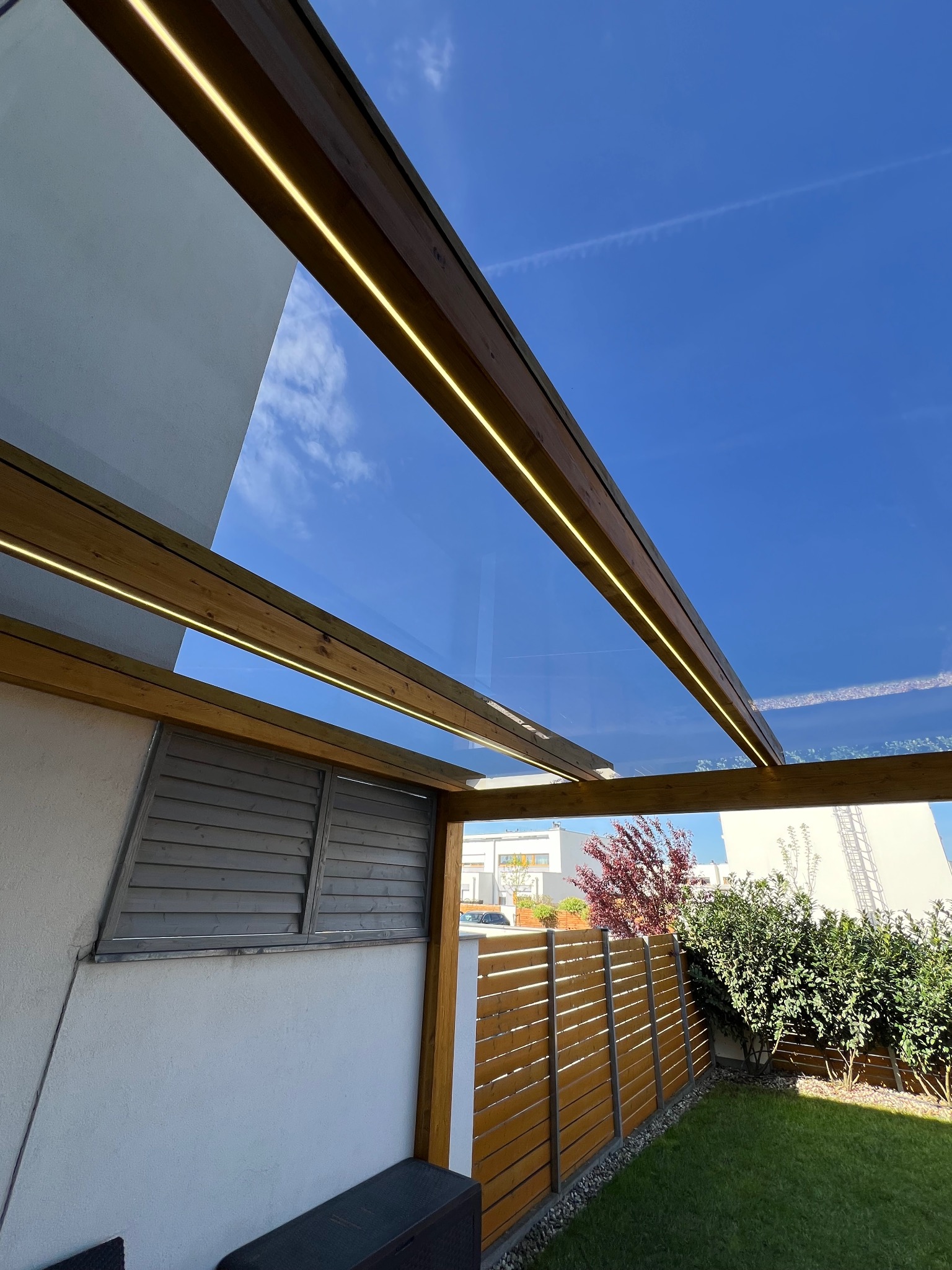 Nowoczesna pergola z drewna z oświetleniem LED i szklanym dachem, widok z dołu na błękitne niebo. Elementy drewniane w ciepłym odcieniu brązu.