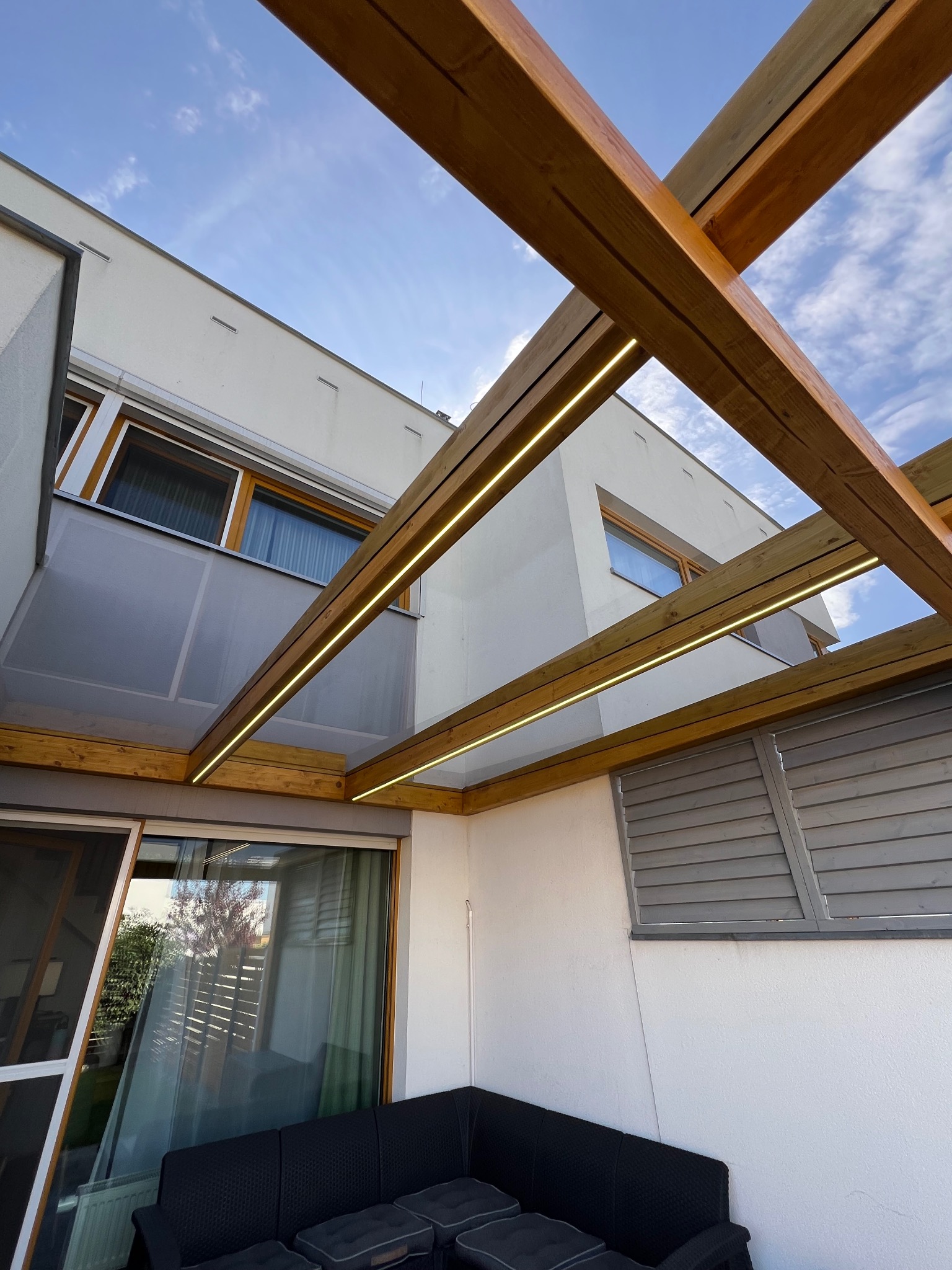 Drewniana pergola z oświetleniem LED w Tarnowie Podgórnym, widok z dołu na konstrukcję i fragment budynku z oknami, błękitne niebo w tle.