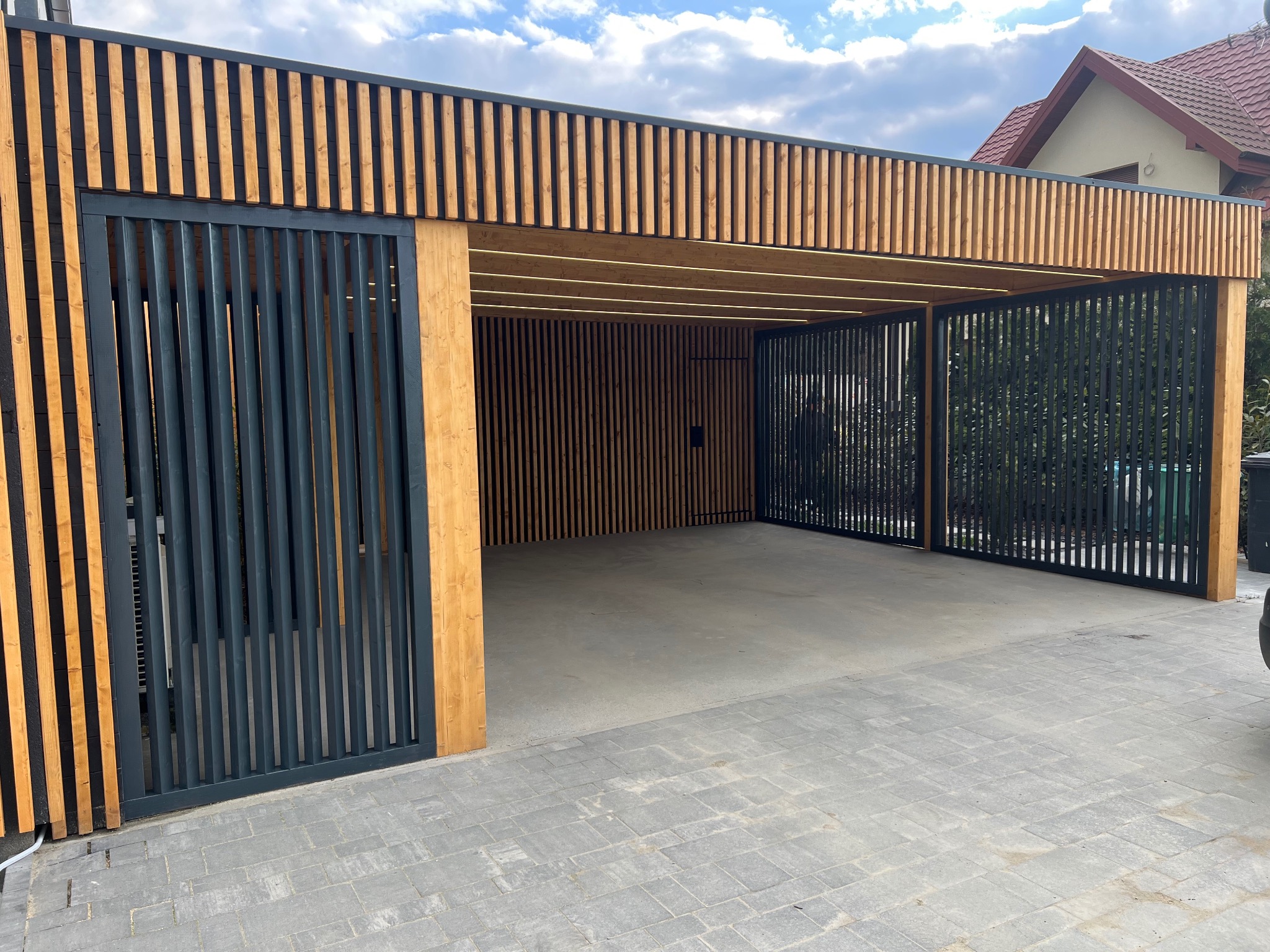 Nowoczesna konstrukcja z drewna i metalu, prawdopodobnie pergola lub wiata garażowa, z elementami pionowych listew i przesuwnymi bramami w odcieniach szarości.