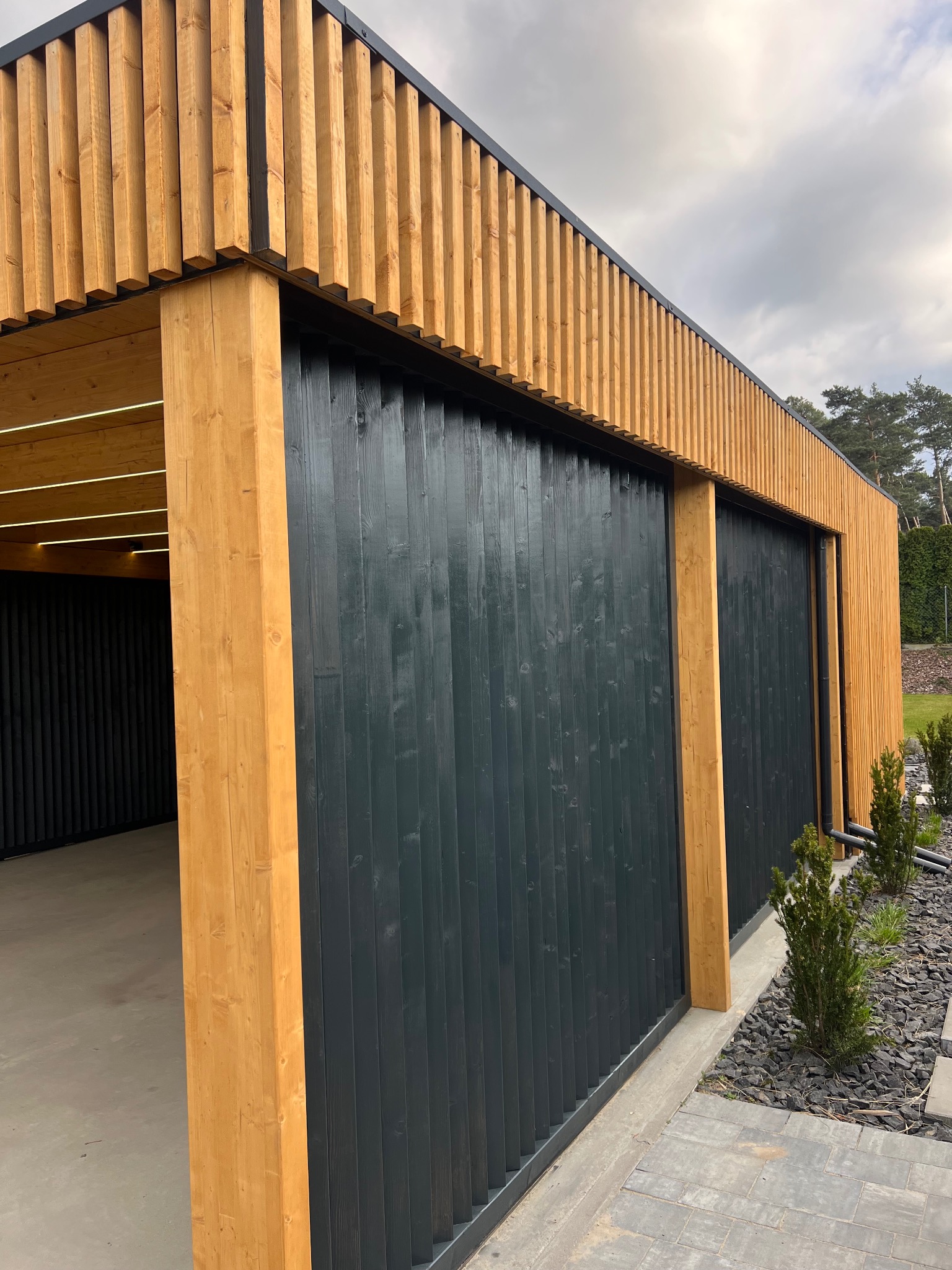Nowoczesna, drewniana konstrukcja z czarnymi, pionowymi panelami i oświetleniem LED, z widokiem na zadbany ogród i szarą kostkę brukową.